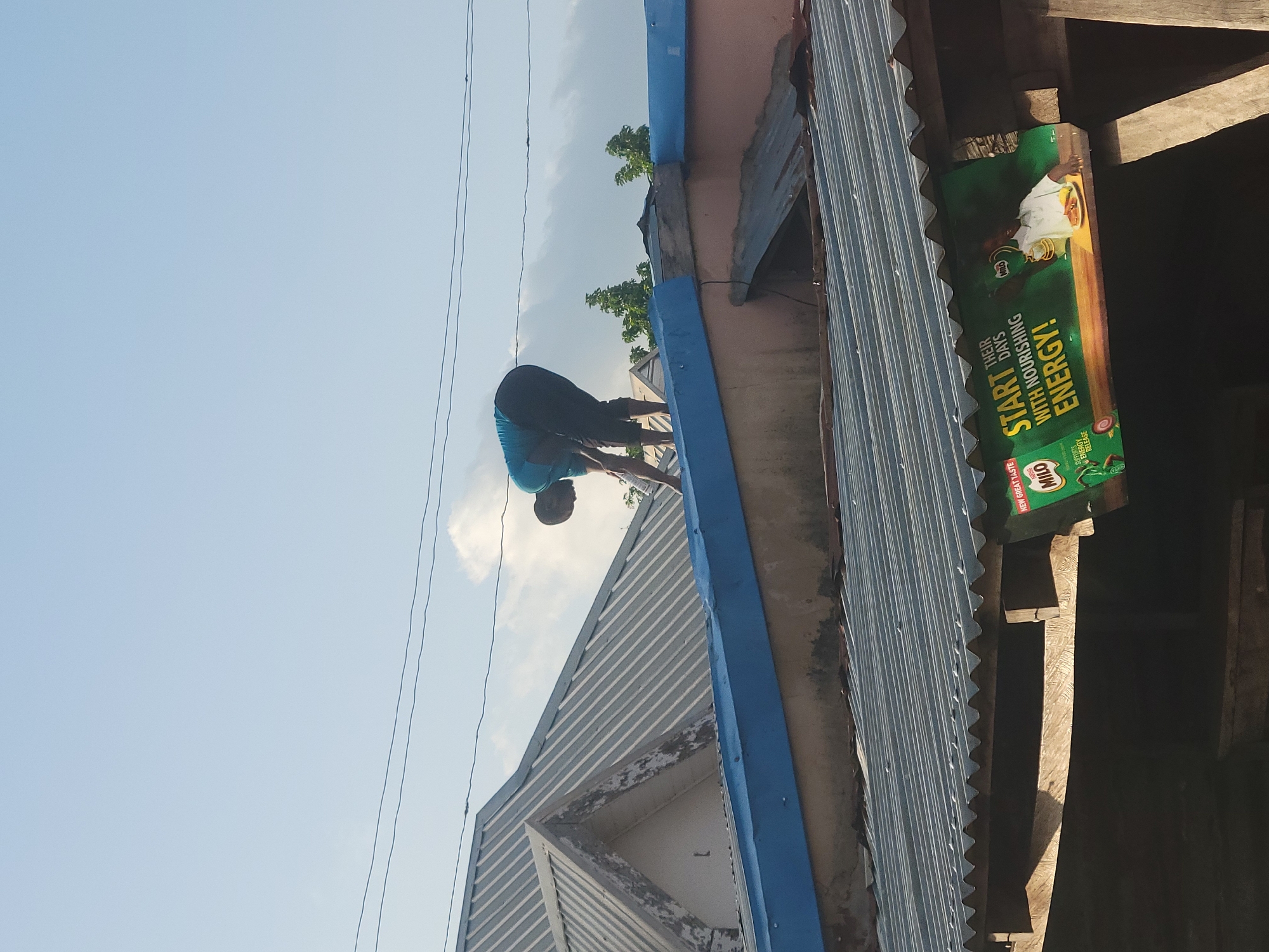 This image captures an outdoor scene in Jos, Nigeria, depicting rooftops and a person engaged in an activity on a building. The sky is light blue with scattered white clouds, indicating daylight. Two dark overhead wires traverse the upper third of the frame.
In the mid-ground, a person is positioned on a flat, blue-painted section of a building's upper facade or parapet wall. They are wearing a blue t-shirt and dark shorts, bending forward significantly with their head down, appearing to perform manual work or inspection on the surface in front of them. Their hands are close to the blue structure. Behind this person and to the left, a multi-gabled, light grey corrugated metal roof with distinct ridges is visible, belonging to an adjacent building. To the right of the person, green foliage from trees can be seen.
In the foreground, a dark grey corrugated metal roof with visible undulations covers a lower structure. Below this roof, supported by wooden beams, a horizontal green advertisement banner for "Milo" is displayed. The banner features text that reads "START THEIR DAYS WITH NOURISHING ENERGY!" and depicts a child holding a cup of Milo with a plate of food. The lower section of the image shows dark, open space beneath this structure, possibly an entrance or stall.