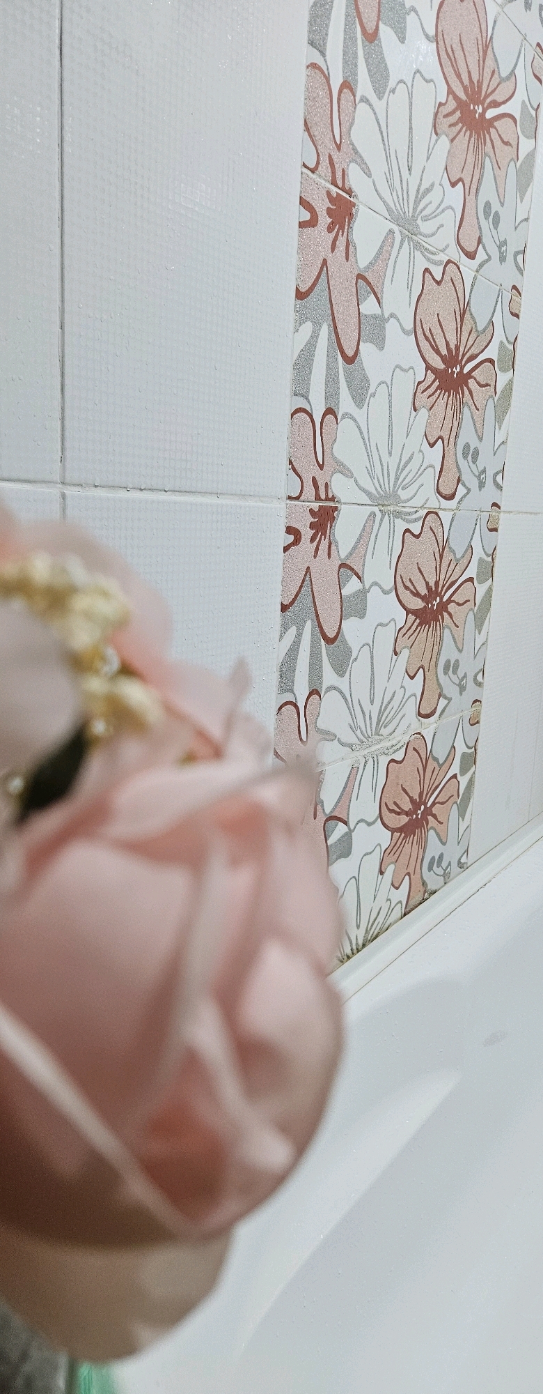 The image shows a close-up of a white tiled wall in what appears to be a bathroom, with a decorative strip of tiles featuring a floral pattern in shades of pink and grey. In the foreground, out of focus, are the soft pink petals of what appears to be a fabric flower, possibly part of a decorative arrangement. A white bathtub edge is visible in the lower part of the image. There are no people present in the image and no identifiable landmarks or features that would indicate the location in Oskemen, Kazakhstan.