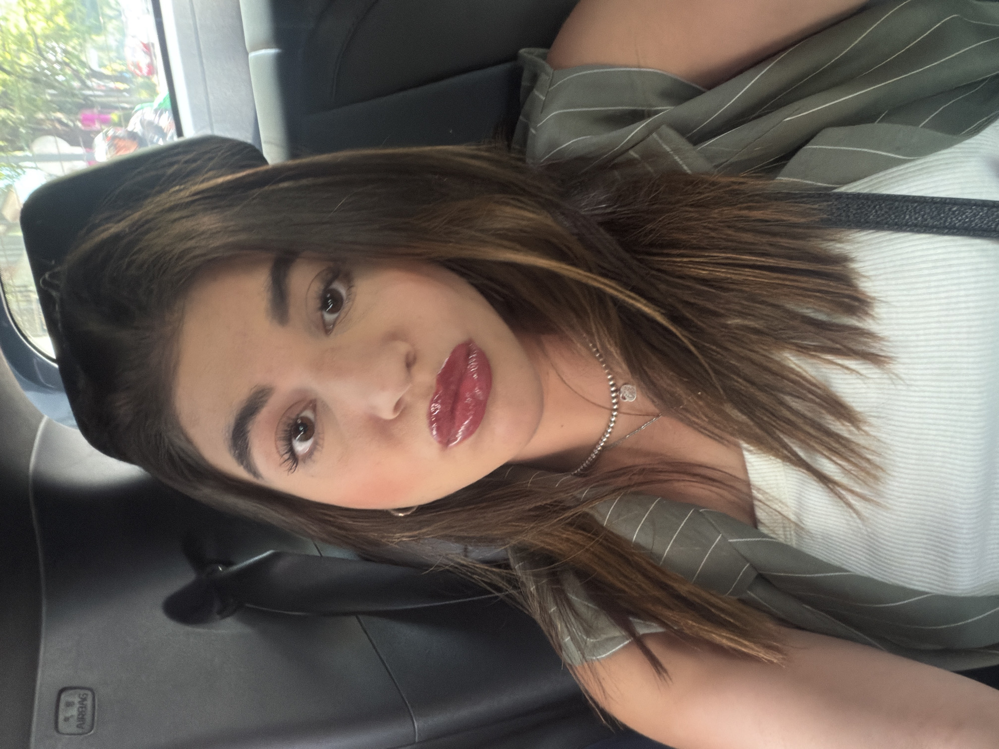 A woman is seated inside a car, likely in the back seat. The car appears to be in motion, as suggested by the blurred background visible through the window. The woman is facing the camera with a slightly tilted head. She has brown hair with highlights, light skin, and glossy lips. She wears a white top underneath a striped, sage green jacket. A silver necklace adorns her neck. Inside the car, one can see the black headrest of a seat and the interior of the vehicle. The reflection in the window may indicate a location in Mexico City, Mexico.