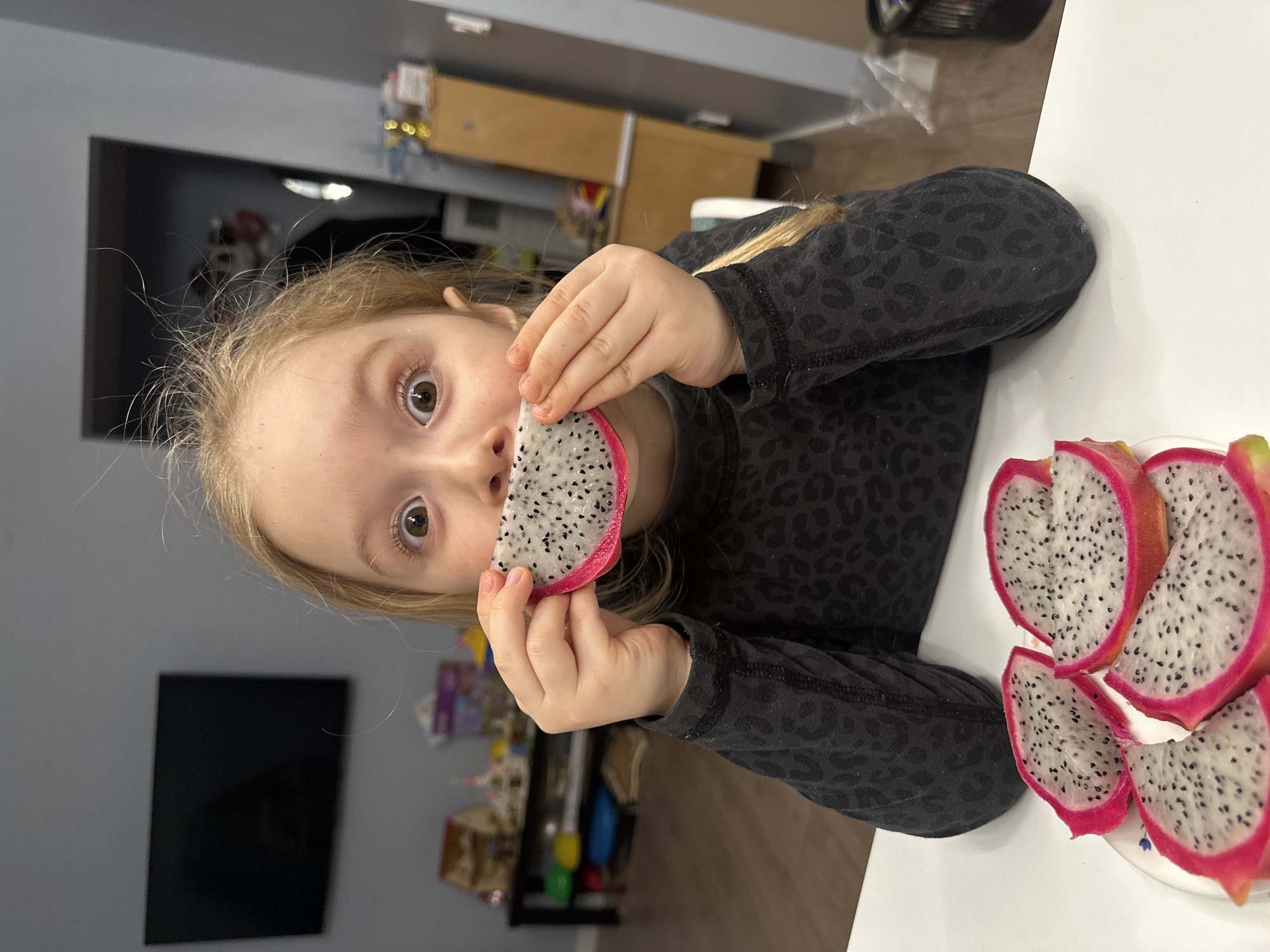 A young, fair-skinned girl with blonde hair is holding a slice of dragon fruit up to her mouth. She has sparkly makeup on her forehead and her eyes are wide open, looking at the camera. She is wearing a dark gray long-sleeved shirt with a leopard print design. A plate of sliced dragon fruit is to her right on a white surface that appears to be a table. The background shows a cluttered interior space with a television, cabinets, and various items. The image was not taken in the city of Chaikovsky, Russia.