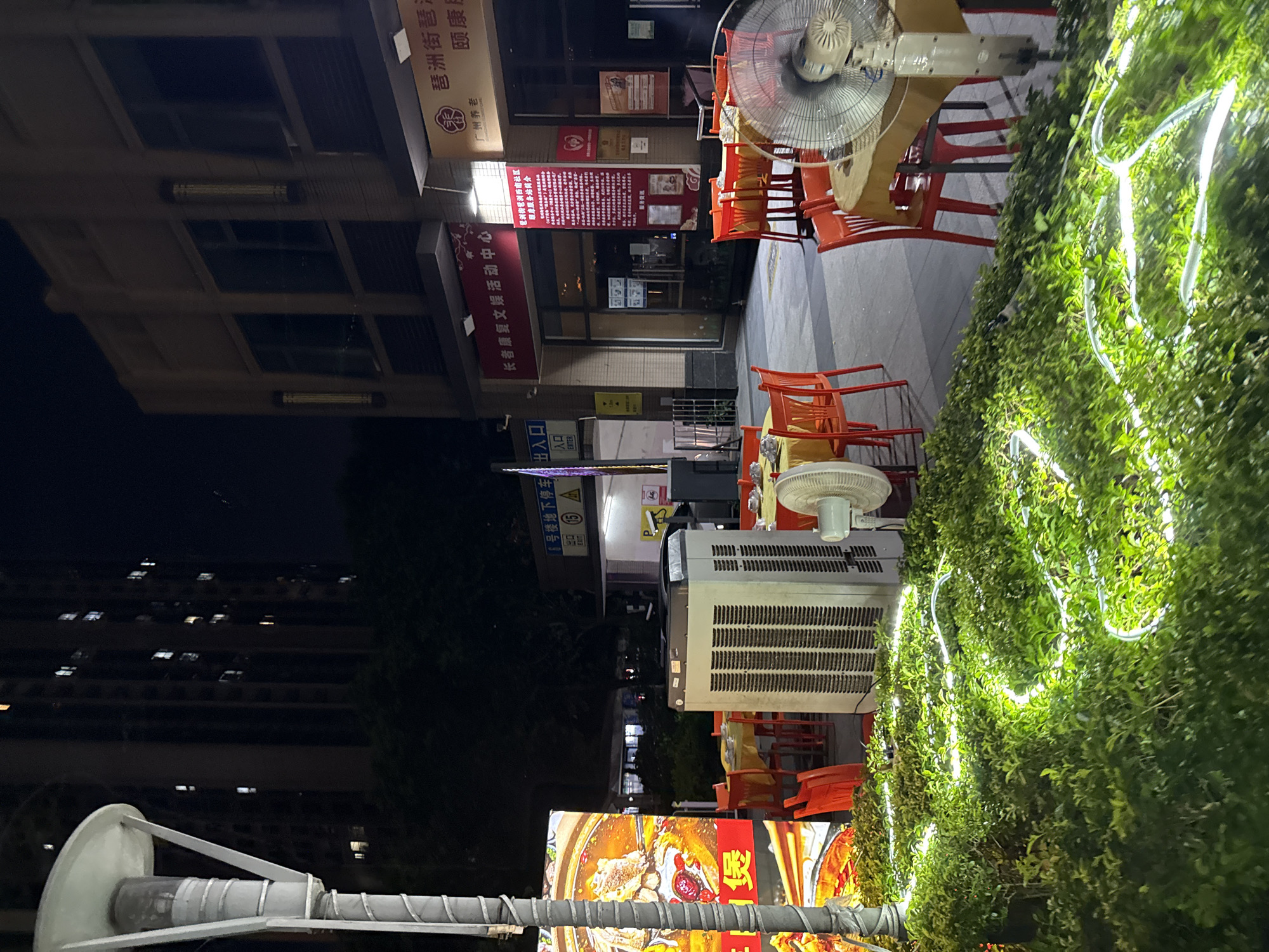 The image depicts an outdoor seating area of a restaurant at night in Haizhu District, China. Red plastic chairs and yellow-clothed tables are set up on a paved surface, suggesting a dining establishment. In the foreground, a well-lit hedge is decorated with white LED strip lights, creating a luminous serpentine pattern. To the left, a large digital billboard displays an advertisement for food, featuring a close-up of a steaming dish. A tall lamppost stands next to the billboard. In the midground, a large white air conditioning unit is positioned between tables, and a pedestal fan stands to the right, indicating an effort to cool the outdoor space. In the background, the facade of a building is visible, with illuminated windows suggesting residential or commercial spaces above. Signs with Chinese characters are present on the building, one of which appears to be a banner for a community activity center. The overall scene is illuminated by artificial lights from the building, the billboard, and the decorative lighting on the foliage, creating a nighttime urban ambiance.