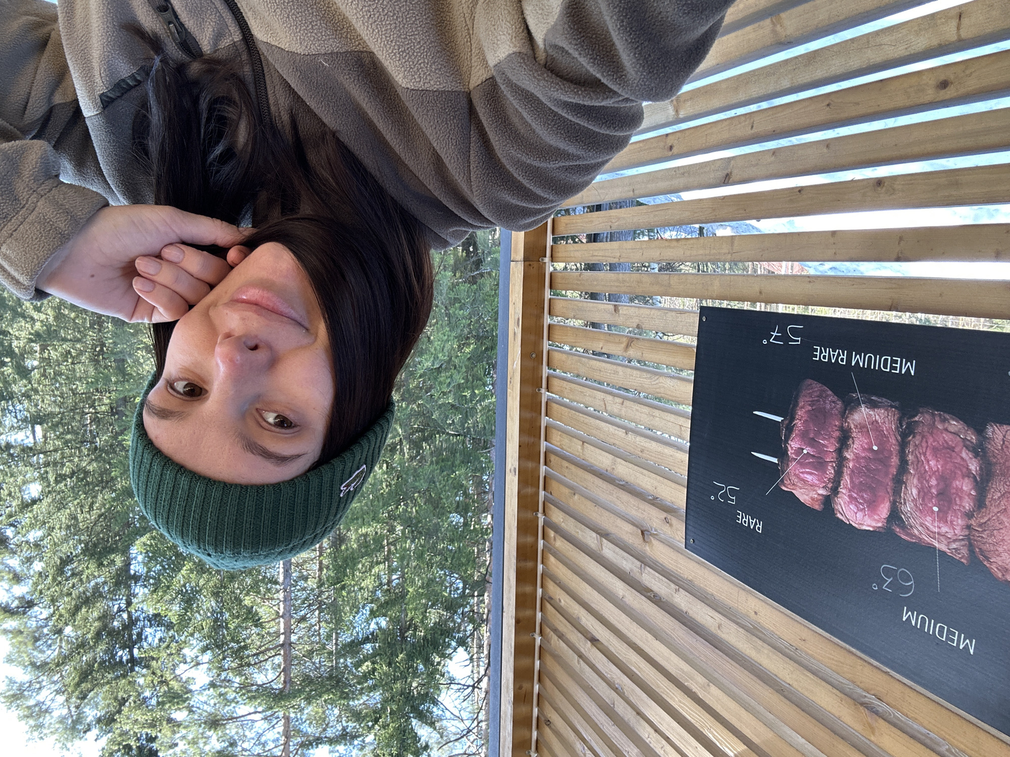 The image depicts an adult female engaged in a selfie within a semi-enclosed wooden structure in Прикамский, Russia. She has dark hair and is wearing a dark green ribbed beanie with a white emblem, paired with a two-tone fleece jacket, primarily taupe with lighter and darker sections, featuring a zipper closure. Her left hand, with manicured nails, rests near her chin as she looks towards the camera with a slight smile. To her left, a black sign affixed to the wooden wall displays an image of various cuts of meat on skewers, accompanied by text detailing doneness levels and temperatures: "MEDIUM 63°", "RARE 52°", and "MEDIUM RARE 57°". The structure features horizontal wooden slats, through which a snow-covered ground and a dense coniferous forest with evergreen trees are visible under a clear blue sky, indicating an outdoor or nature-adjacent setting.