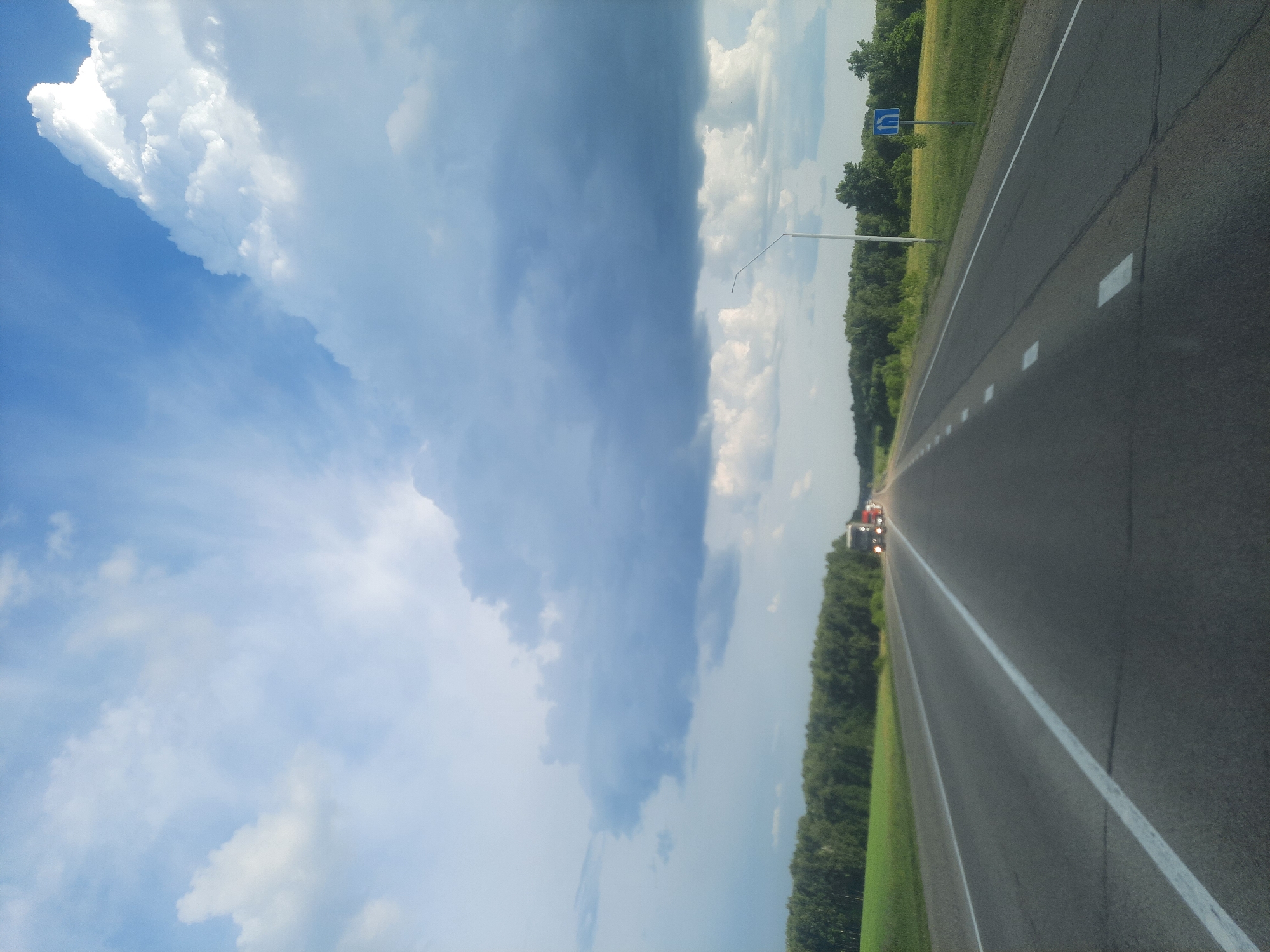 A forward-facing, eye-level, long shot captures a highway in daylight, likely in the city of Oktyabrsky, Russia. The asphalt road stretches into the distance, marked with solid white and dashed white lines. Two lanes are visible in the foreground, with what appears to be a breakdown lane on the right.
On the right side of the road, a green field is bordered by a dense line of dark green trees. A single white lamppost stands prominently in the mid-ground. Further down the road, a blue sign with white arrows indicating a lane merge is visible. On the left side of the road, a bright green field meets the tree line.
In the distance, several trucks are traveling on the highway, with their headlights illuminated. The sky above is filled with dramatic clouds, ranging from bright white cumulus formations to darker, more ominous storm clouds. The overall lighting suggests either early morning or late afternoon.