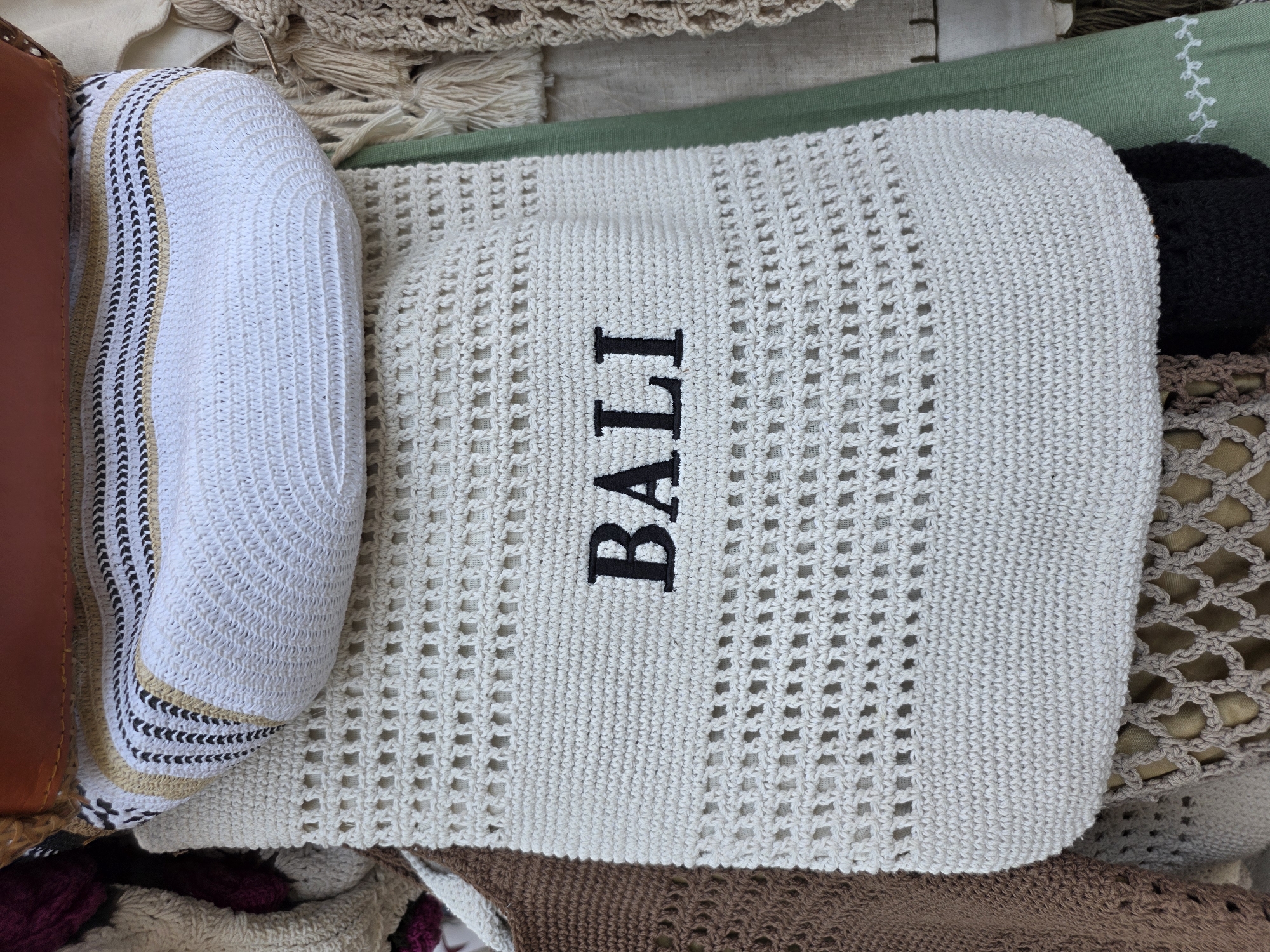 A close-up, high-angle shot shows a cream-colored, crocheted tote bag with "BALI" embroidered in black capital letters. The bag has a patterned opening with a brown leather strap at the top. It is part of a display of handcrafted items. The background is blurred, but suggests other textile goods, possibly in a market stall. The scene is indoors or shaded, with soft lighting. The location is identified as North Kuta, Indonesia.