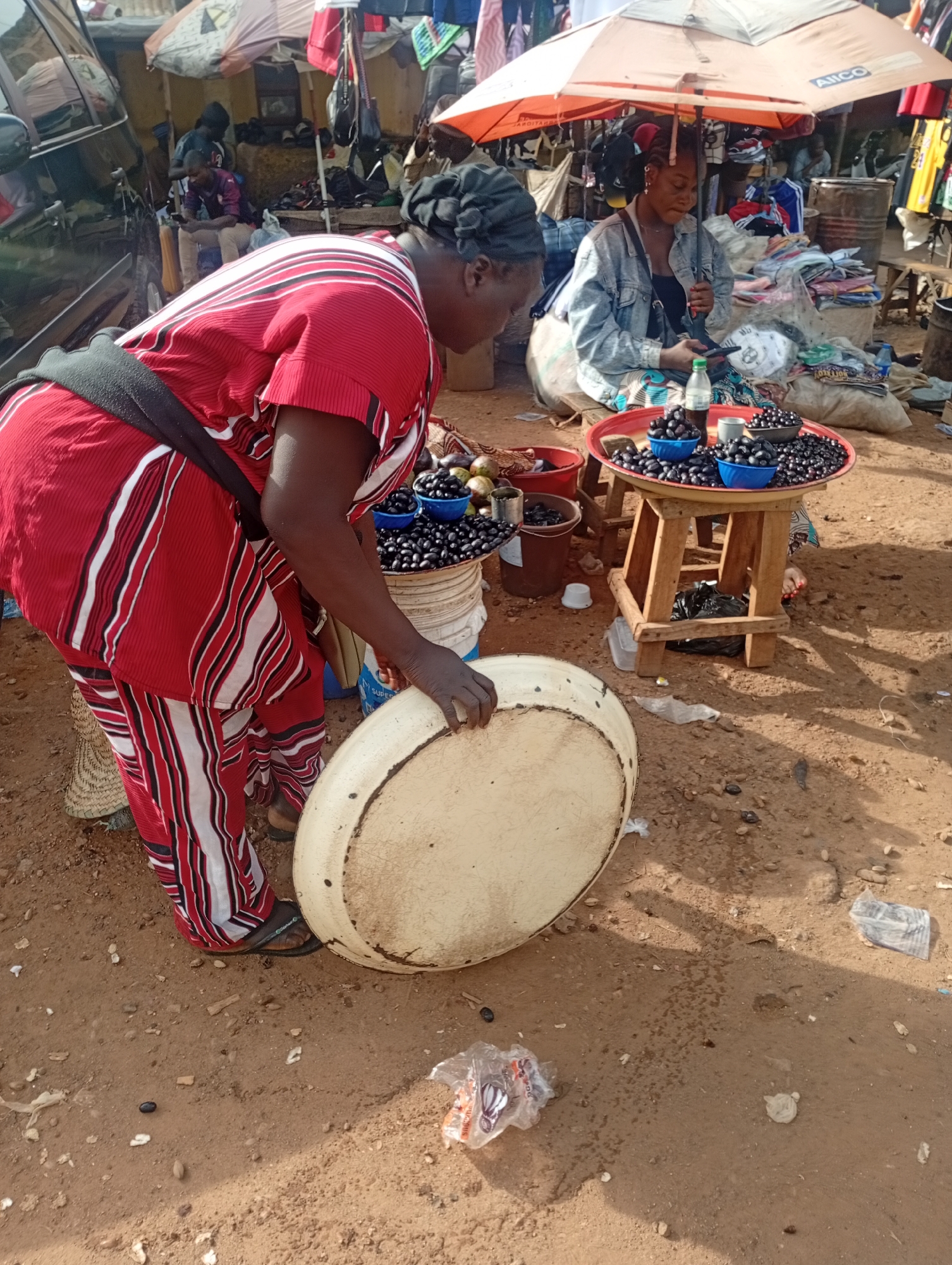 This media file captures a scene in an outdoor market in Rafin Par, Nigeria. The primary subject is a woman in the foreground, dressed in a red and white striped outfit and a black sash, who is bent over, holding a large, light-colored circular object. Her attention is focused downwards.
Behind her, a second woman sits under a bright orange umbrella that displays the text "AJICO". This woman is wearing a denim jacket over a black top and is looking at her phone. A table with wooden legs sits in front of her, laden with a red tray holding several blue bowls filled with dark, round fruits, likely berries. Bottles of liquid are also visible on the tray.
The setting appears to be a busy marketplace with stalls and various goods on display, including clothing and what seem to be other fruits or produce in the background. There are also barrels and buckets visible, along with a black vehicle on the left side of the image. The ground is covered in dry, sandy earth, and there are scattered plastic bags and debris, suggesting a typical market environment. The lighting indicates it is daytime, likely with bright sunlight. The overall impression is one of a bustling commercial activity.