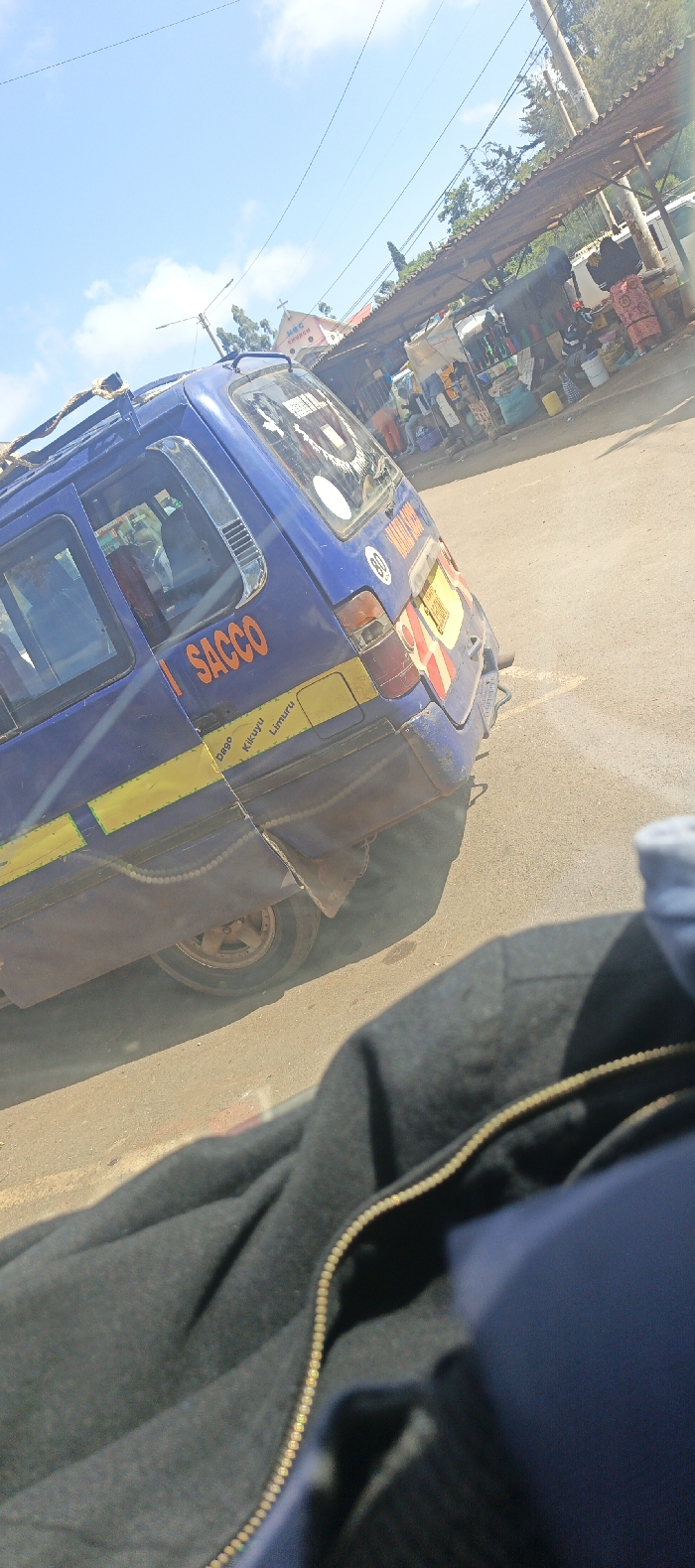The image captures a vibrant street scene, likely in an urban or semi-urban area, during bright daylight hours. The perspective is from a slightly elevated position, possibly from inside another vehicle, looking out.
In the foreground, the bottom right corner is occupied by a dark, textured fabric, resembling a bag or clothing, featuring a prominent golden-colored zipper running diagonally upwards.
The central subject of the image is the rear and side of a blue public transport van, commonly known as a matatu in East Africa. It has yellow horizontal stripes along its lower body. Orange lettering on its side clearly reads "SACCO," indicating its affiliation with a cooperative society. Below the side windows, on the yellow stripe, smaller text specifies destinations or routes: "Dego Kikuyu Limuru," which are towns in Kenya. The rear of the van shows a partially visible yellow license plate area with red markings, and a circular white sticker with the number "80," likely indicating a speed limit. The vehicle appears well-used, with some dirt or dust visible on its body and the rear wheel. A rope or tie-down system is visible on the roof rack.
In the background, beyond the blue van, a lively market area unfolds. There are several stalls with corrugated iron roofs, underneath which various goods are displayed for sale. Several individuals, likely vendors or shoppers, can be seen near these stalls. Further into the background, to the left of the market stalls, a building with a light-colored facade and a red roof is visible. A cross adorns its peak, identifying it as a church. The visible text on the church building confirms it is "M.A.C CHURCH."
The sky above is bright blue with scattered white clouds, suggesting clear weather. Multiple power lines crisscross the upper part of the sky. Green foliage and trees are visible behind the market stalls and church, indicating some natural elements in the urban landscape. The ground appears to be a dusty road or unpaved street.