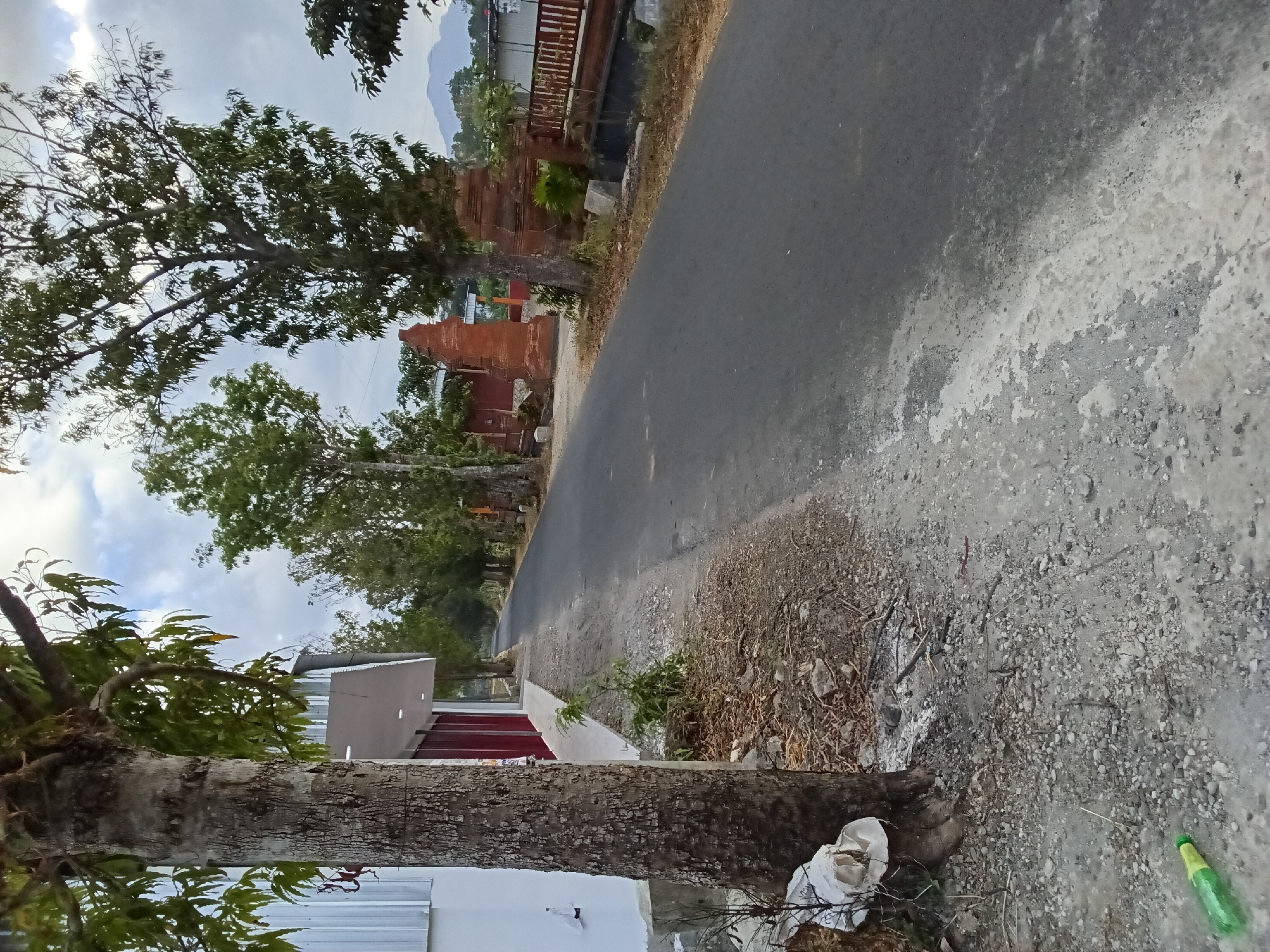 The image depicts a paved road in Bendorejo, Indonesia, with a sidewalk on the left and trees lining the right. A building with a white facade and red doors is visible on the left, adjacent to a large tree trunk. The road continues into the distance, flanked by more trees. In the mid-ground, there are traditional brick gates, a red-painted fence, and a small canal. In the background, hills can be seen under a cloudy sky. The ground along the sidewalk is covered in gravel and dry leaves. A green plastic bottle lies on the ground in the lower left corner. There are no people visible in the image.