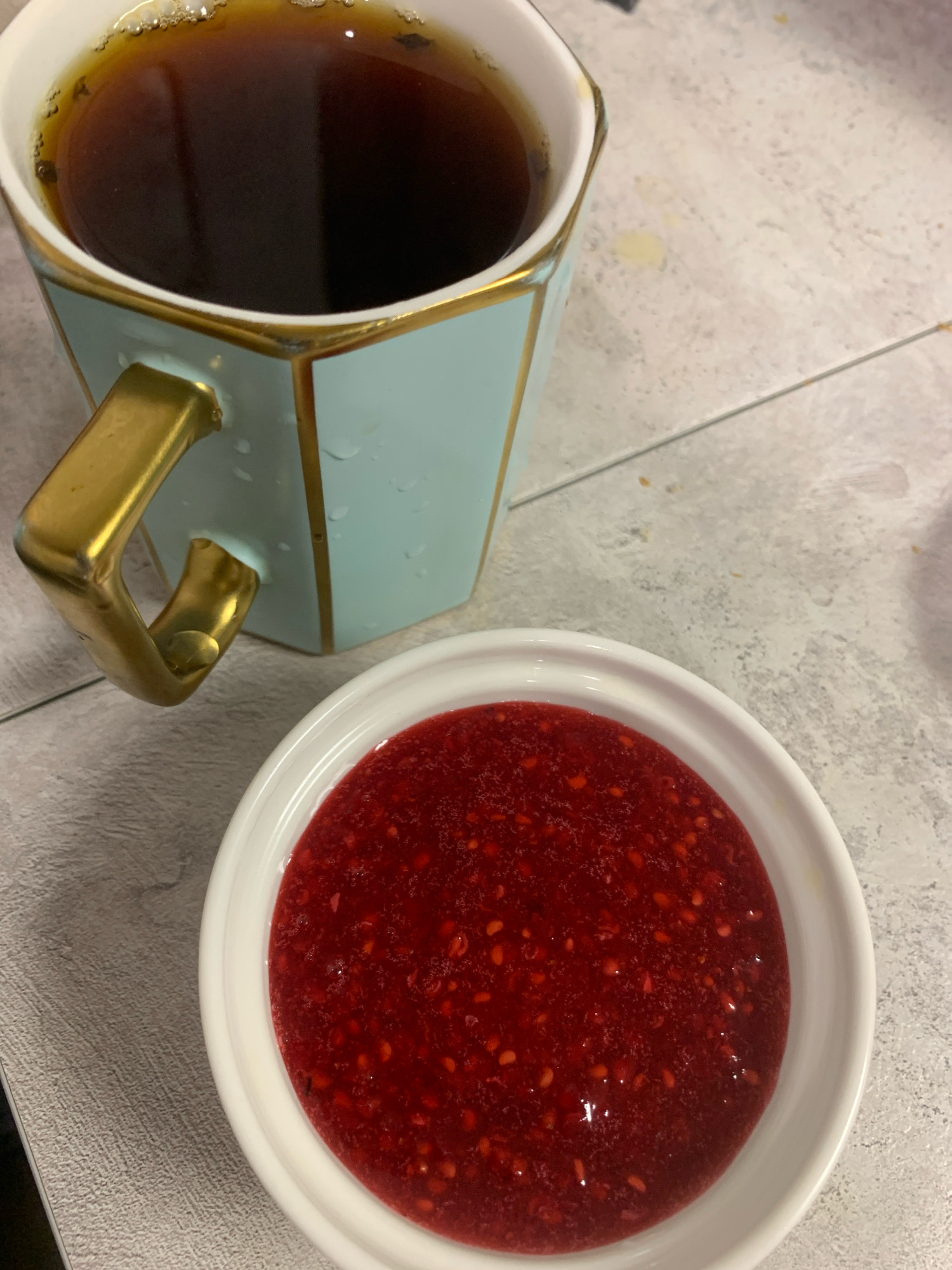 A close-up shot shows a light blue mug with a gold handle and gold trim filled with dark liquid, likely coffee or tea, on a textured, light-colored surface. Next to the mug is a white bowl filled with a vibrant red, textured substance, appearing to be raspberry jam or compote. The mug has water droplets on its exterior, and the surface it rests on has a subtle pattern and a few faint yellow or brown smudges. The lighting suggests an indoor setting. The image does not contain any elements that would suggest its location in the city of Kyakhulay, Russia.
