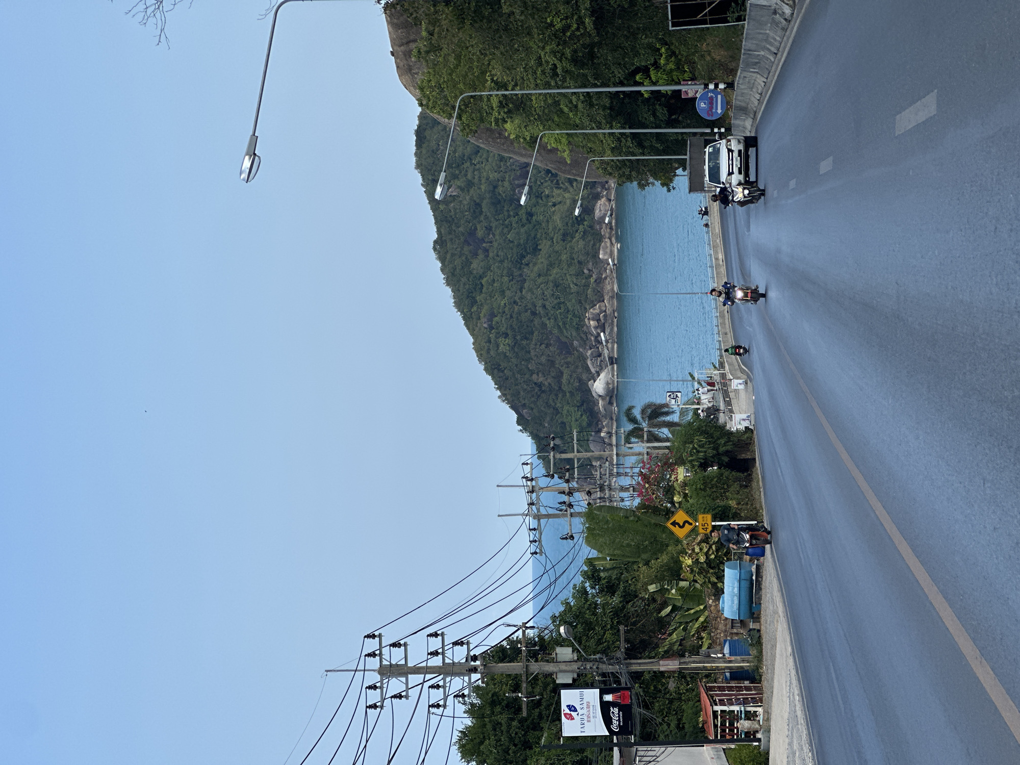 The image captures a vibrant coastal road scene, likely in Baan Chaweng Noi, Thailand, as indicated by the location context and visible Thai script. The perspective is from the side of a paved road, looking ahead as it gently curves.
On the right, the two-lane road features a solid yellow center line and white dashed lane markers. Several vehicles, primarily motorcycles and scooters carrying individuals, are traveling on the road, with a white van also present further ahead. Street lights with extended arms line the road, their reflective surfaces catching the daylight.
To the left, a steep, verdant mountain or hill covered in dense green foliage rises, meeting the tranquil, blue-green waters of the sea. The coastline appears rocky in sections. A prominent feature on the left is a dense array of utility poles and overhead power lines, characteristic of the region, which partially obscure the view of the coastline and some structures. Tropical trees, including palm trees, dot the landscape between the road and the sea.
Visible road signs include a blue circular sign with a white 'P' symbol, indicating parking, and a yellow diamond-shaped warning sign with black wavy arrows, signifying a winding road, accompanied by the text "45 km/h," likely an advisory speed limit. A large billboard or sign is also visible on the left, displaying "TARULA SAMUI" in English, alongside Thai script and a "Coca-Cola" logo.
The sky is a clear, bright blue with no clouds, suggesting good weather conditions during daytime, possibly late morning or early afternoon. A tiny, dark speck is visible high in the sky. The overall scene depicts active transportation along a scenic tropical route.