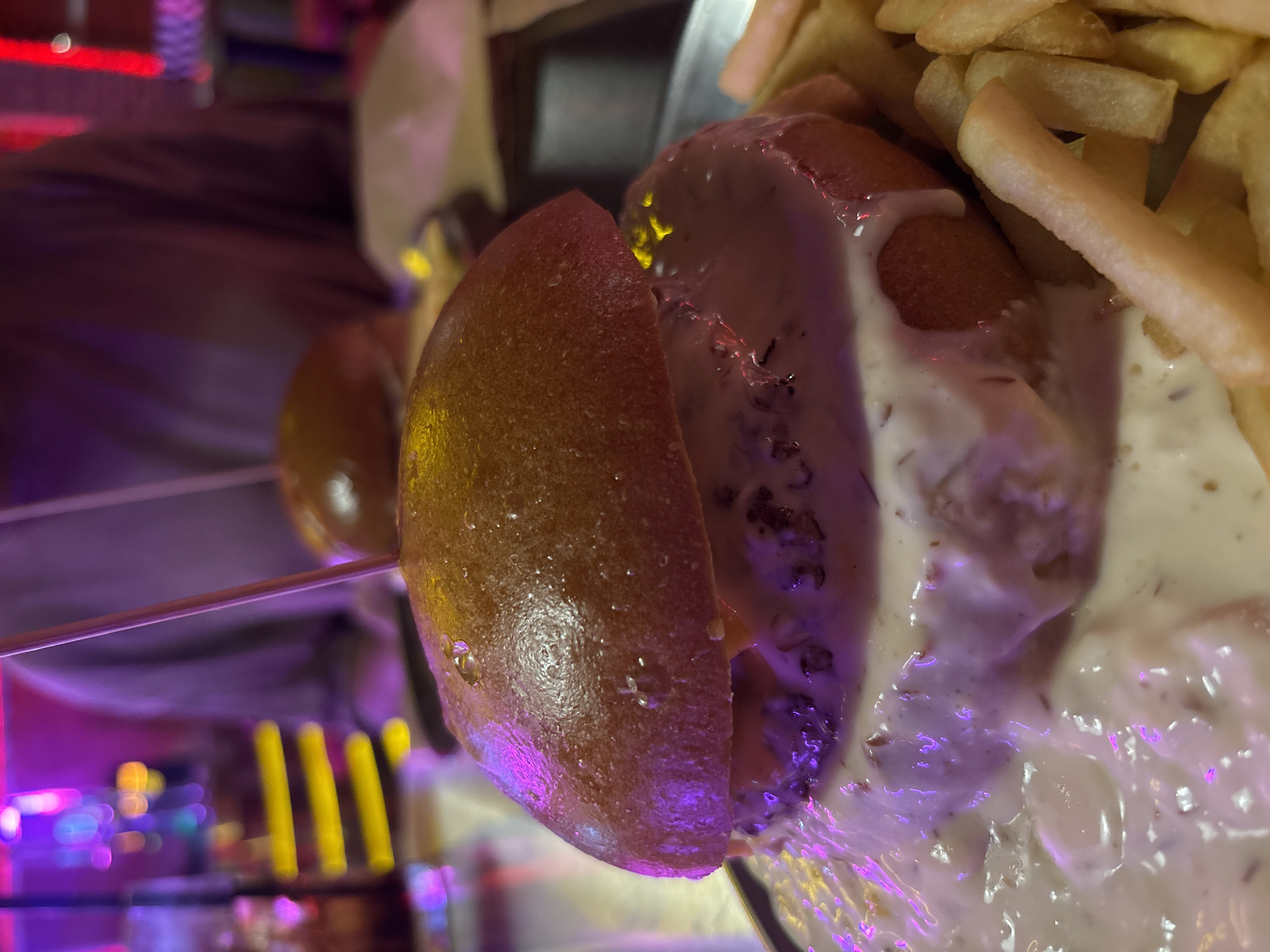 A close-up, low-angle shot captures a large, decadent burger with a thick layer of creamy sauce overflowing from its sides. The burger is served with a generous portion of golden french fries on a metallic plate. In the background, blurred figures are visible, as is a neon-lit establishment, suggesting the setting is a vibrant restaurant or bar. The lighting is dim, with pink and purple hues dominating the scene, creating a moody and atmospheric ambiance.