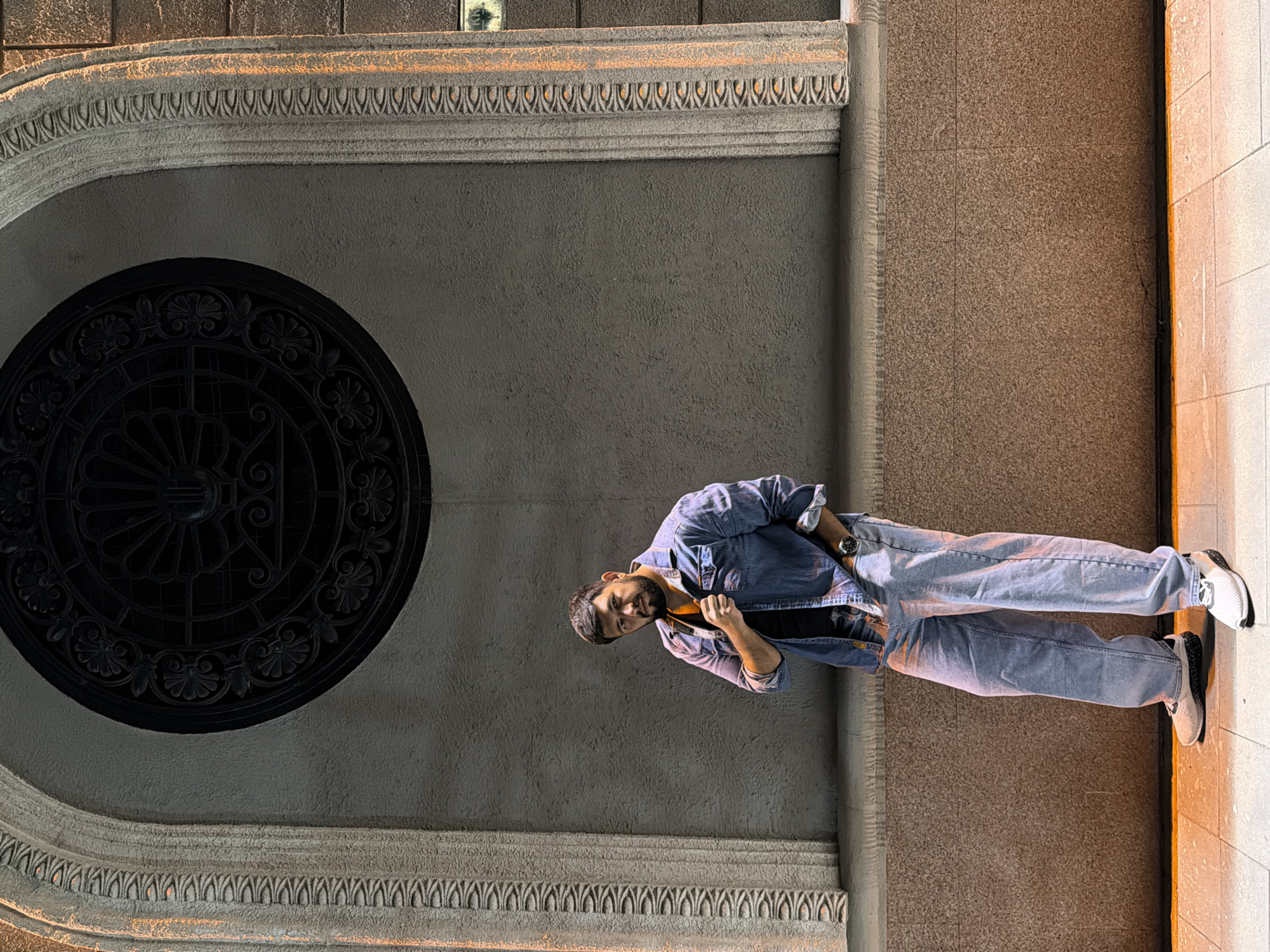 A low-angle shot of a man standing in front of an architectural archway. The man is wearing a denim shirt, a black undershirt, light-wash jeans, and sneakers with black and white soles. The architectural feature behind him has a black circular decorative element. The location appears to be urban, with a tiled wall to the right of the man.