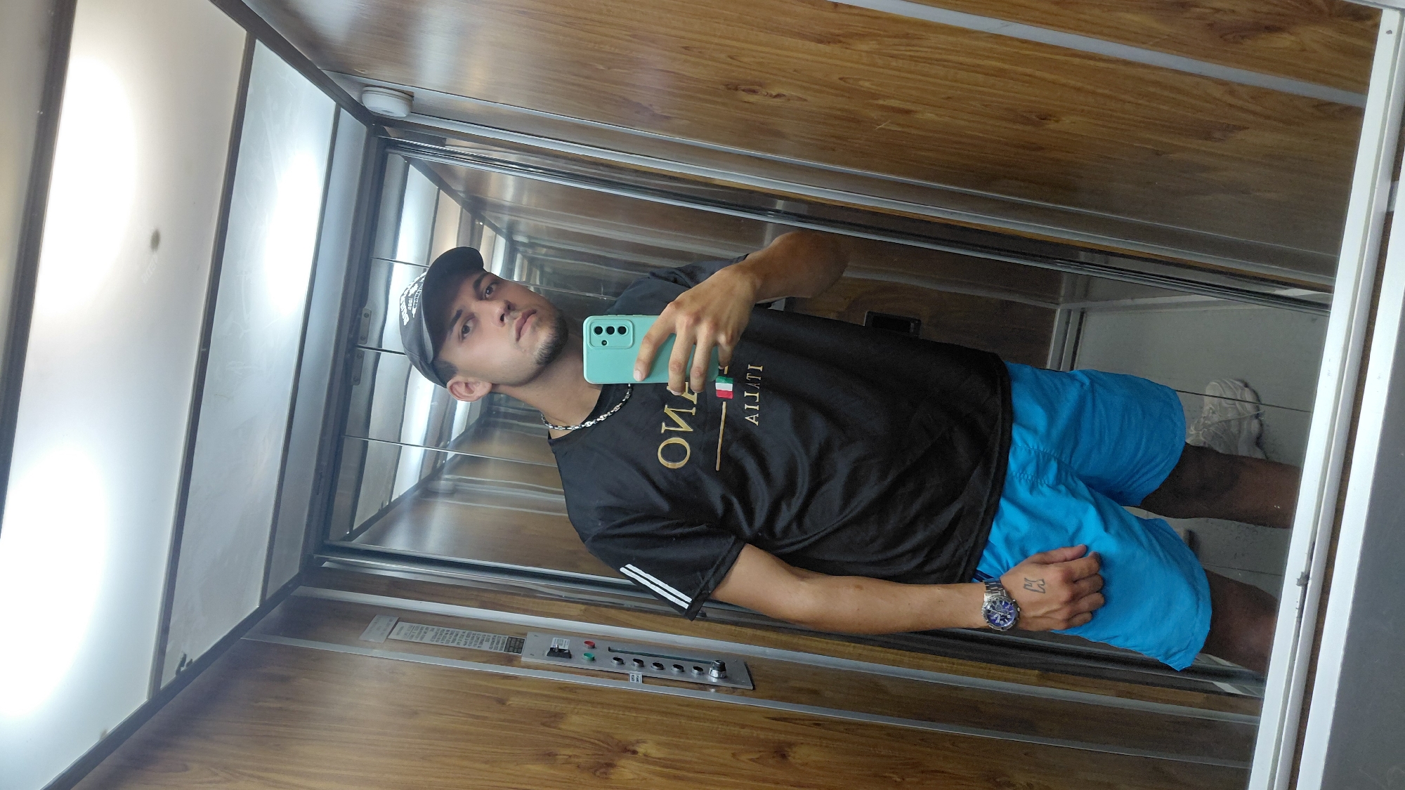 A young man with a goatee is taking a mirror selfie in an elevator. He is wearing a black t-shirt with white stripes on the sleeves and a gold graphic with the word "ITALIA" and the Italian flag, a silver chain, blue shorts, a wristwatch on his left wrist, and a black baseball cap. He is holding a light blue smartphone in his right hand, which is angled to capture the mirror image. The elevator has wood-paneled walls and reflective metal accents. A control panel with buttons is visible on the left wall. The floor of the elevator is grey. The man's left hand has a tattoo of the letter "K" on it. The overall scene suggests a casual, everyday moment captured within an urban elevator.
