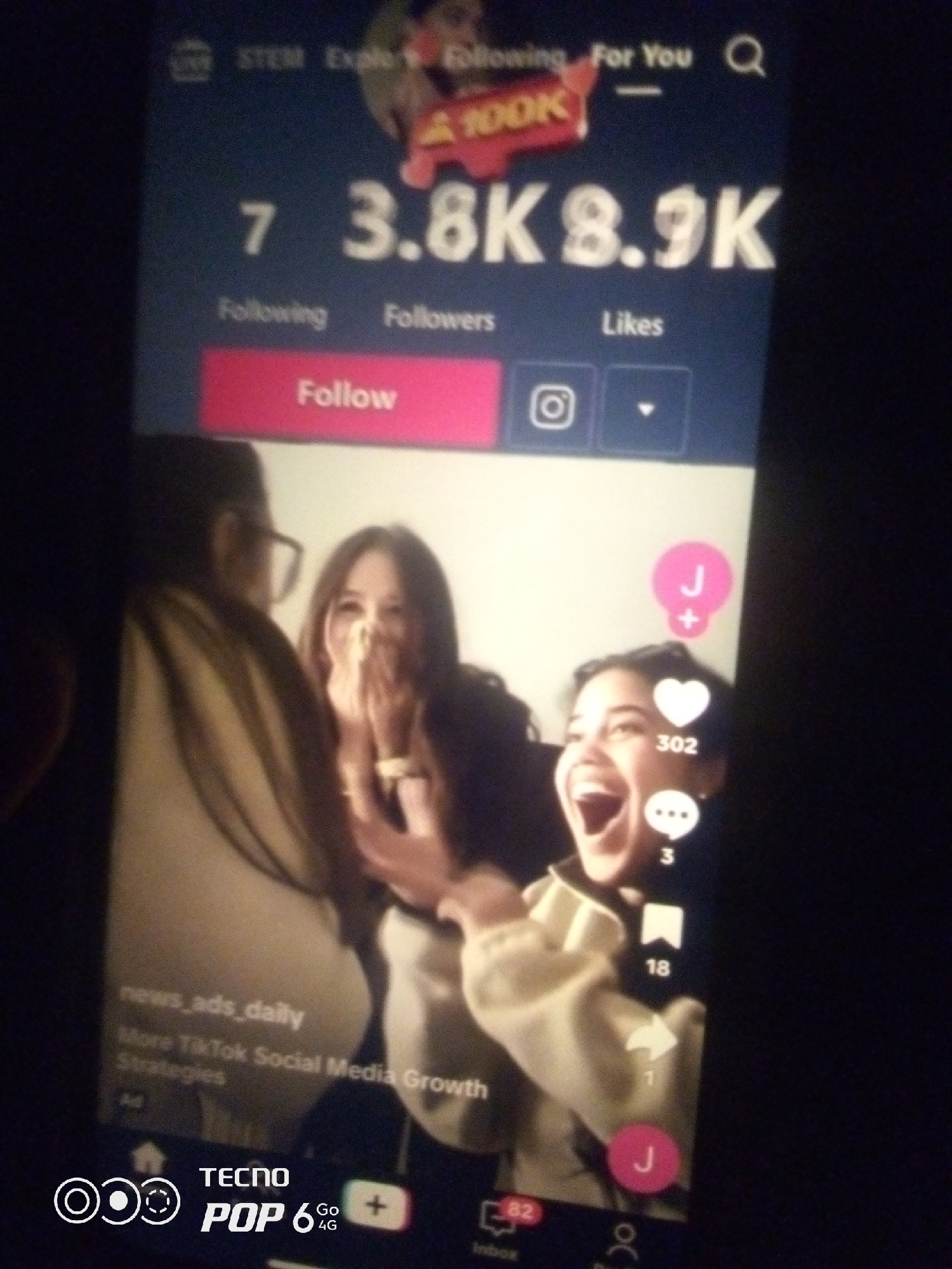 The image displays a smartphone screen with a social media profile page, likely from TikTok, superimposed over a dimly lit background. The screen shows a profile with the username "news_ads_daily" and the description "More TikTok Social Media Growth Strategies." Metrics visible include "7 Following," "3.8K Followers," and "8.9K Likes." A prominent "Follow" button in pink is present, along with icons for Instagram and a down arrow. Below this, a video is playing, featuring three individuals. The person in the foreground is a young woman with an open-mouthed, joyful expression, her hands covering her mouth. To her left, another young woman with glasses is partially visible. To the right of the main subject, a third individual's face is partly seen. Interactive icons are displayed on the right side of the video, including a pink circle with a "J+" inside, a heart icon with "302" below it, a speech bubble icon with "3" below it, a bookmark icon with "18" below it, and a share icon with "1" below it. At the bottom of the screen, a navigation bar is visible with icons for "Inbox" (with "82"), a user profile, and "Home." The brand "TECNO POP 6 4G" is visible in the lower left corner. The image's overall quality is low due to blurriness and poor lighting, suggesting it was taken in low light conditions, potentially indoors. There is no information within the image to suggest its location in Howayi, Nigeria.