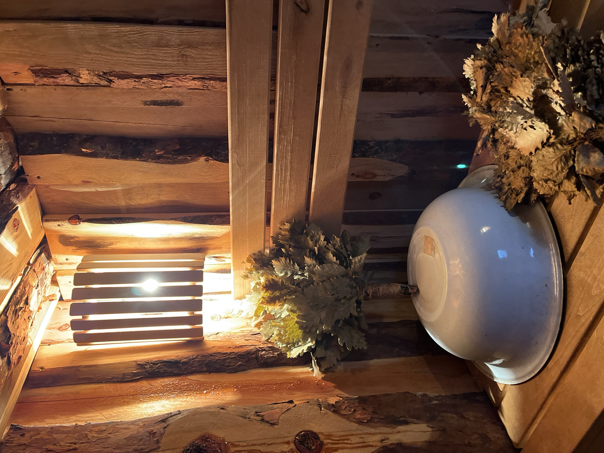 The image depicts the interior of what appears to be a sauna in Зыково, Russia. The walls and ceiling are constructed of rough-hewn logs, some retaining bark. A slatted wooden structure with a light source behind it provides illumination. Hanging on the walls are two bath brooms (bundles of leafy branches) and a white enameled basin. The scene is dimly lit and evokes a sense of rustic functionality.