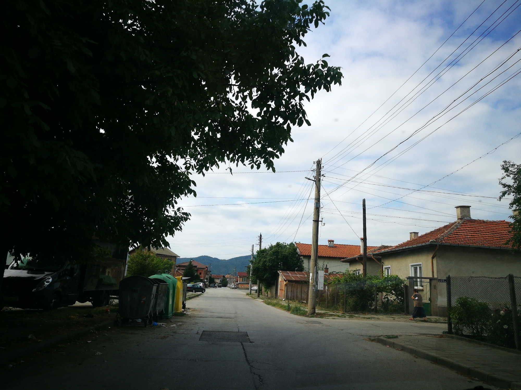 The image shows a street scene in Ihtiman, Bulgaria. The street is paved and appears to be a residential area. There are houses with red tile roofs on both sides of the street. A person is walking on the sidewalk in front of one of the houses. There are several garbage bins on the side of the road, and a truck is parked nearby. Overhead, there are power lines strung between poles. In the background, hills are visible under a cloudy sky. The overall impression is a somewhat weathered but lived-in urban environment.