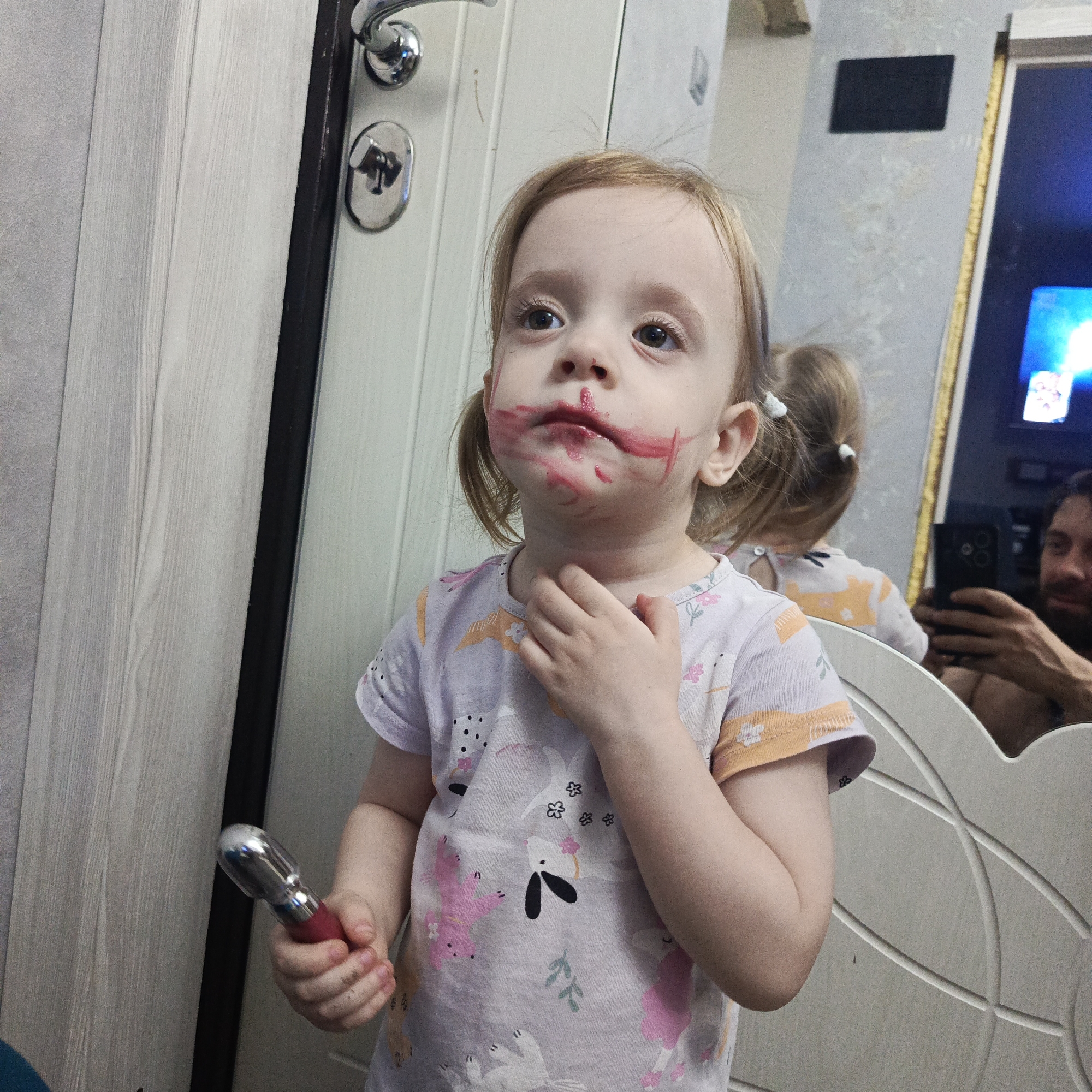 A young, fair-skinned child with reddish-blonde hair styled in pigtails stands in front of a mirror. The child's face is smeared with red lipstick around the mouth and cheeks. The child is wearing a light-colored t-shirt with floral and animal designs and holding a red and silver object in their left hand. The child is touching their neck with their right hand.
In the reflection of the mirror, a man is visible holding a phone, possibly taking the picture. A television screen is also visible in the mirror's reflection. The room contains a white door with a silver handle.
The image was taken in Марковский, Russia.
