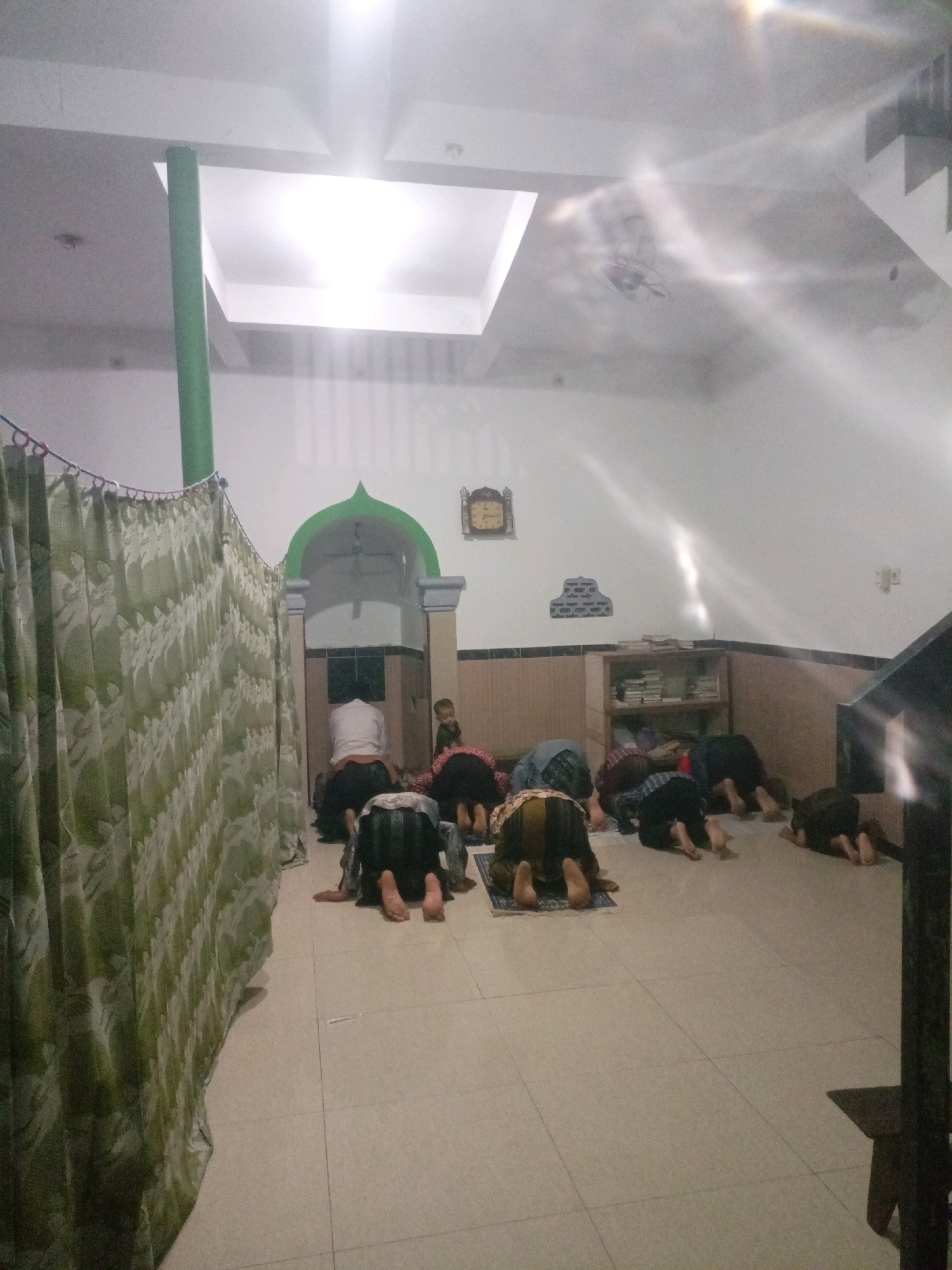 In Manggar, Indonesia, a group of people are engaged in prayer inside a room that appears to be a place of worship, possibly a mosque or a prayer hall. The room has white walls and a tiled floor. A green curtain with a leaf pattern is drawn across the left side of the image, separating an area from the main prayer space. Several individuals, dressed in traditional Islamic attire, are positioned on prayer mats, bowed in supplication. The lighting in the room is bright, with a skylight visible in the ceiling. In the background, an arched doorway with green trim leads to another area, where a clock is visible on the wall. A small child stands near the doorway. A shelf unit filled with books is also present in the background. The overall scene depicts a moment of religious devotion within an indoor community space.
