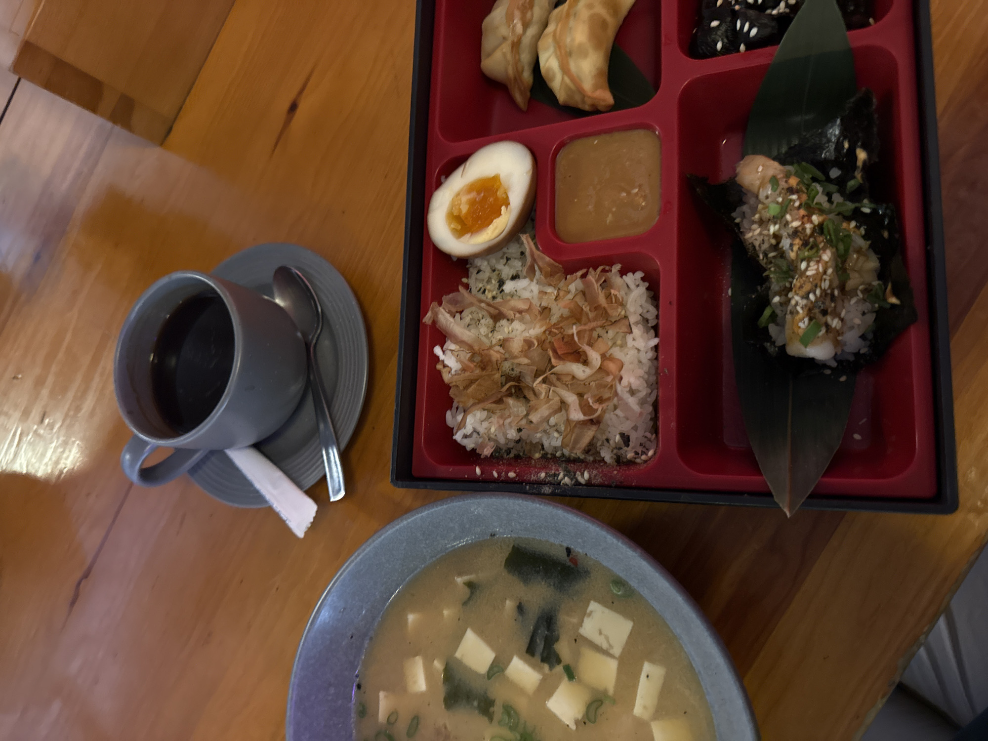 The image shows a table with a meal and a drink. There is a bento box containing gyoza, a boiled egg, a sauce, rice with bonito flakes, and a seaweed wrap with rice and sesame seeds. Next to the bento box is a cup of coffee with a spoon and a sugar packet on a saucer, and a bowl of miso soup. The meal is set on a wooden table. The location is unknown and cannot be determined from the image.