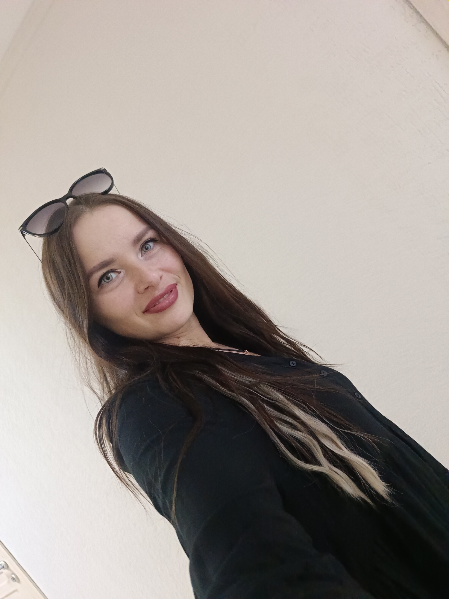 A woman with long brown hair and sunglasses perched on her head is facing the camera. She has fair skin, blue eyes, and is wearing a dark-colored button-up shirt. Her hair has a few blonde highlights. The background is a plain white wall. The location is specified as Chaikovsky, Russia.