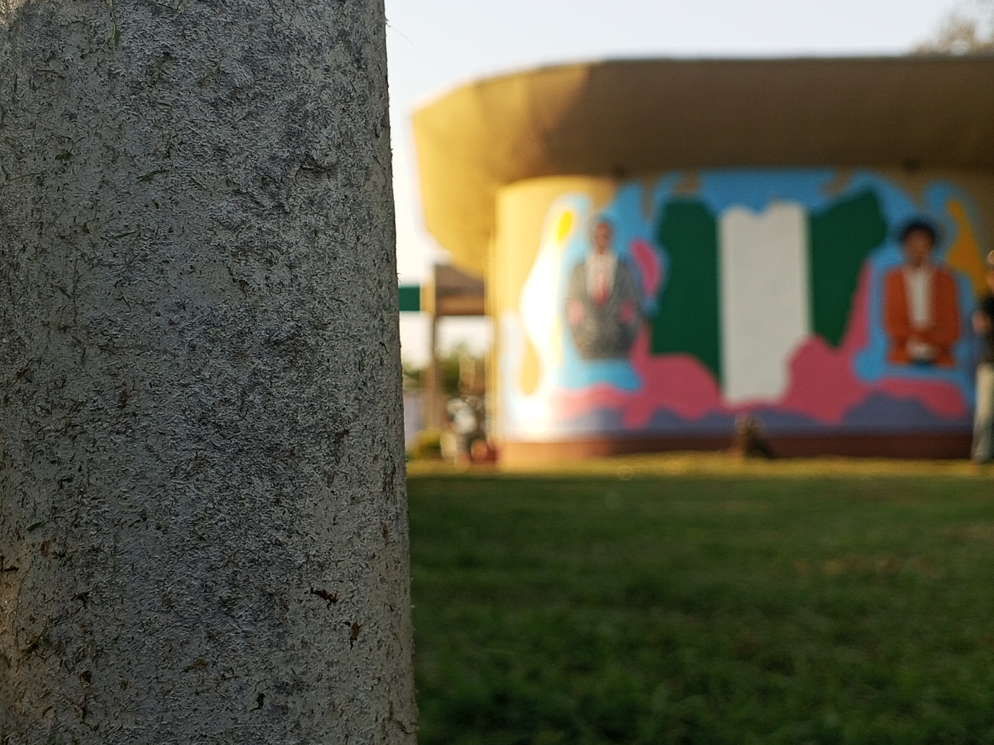 A medium shot captures a cylindrical object, possibly a tree trunk, in the foreground with a rough, textured surface. In the background, slightly blurred, is a building with a broad, overhanging roof. The building's exterior is painted with colorful, abstract designs, including figures resembling human portraits and what appears to be the Nigerian flag. A grassy field extends from the foreground towards the building. Other elements are partially visible, including a structure reminiscent of a bus stop or covered area, along with a person standing near the right edge of the frame. Based on the description, the location appears to be in the city of Tawpo, Nigeria, as indicated by the potential presence of the Nigerian flag and the specification provided.
