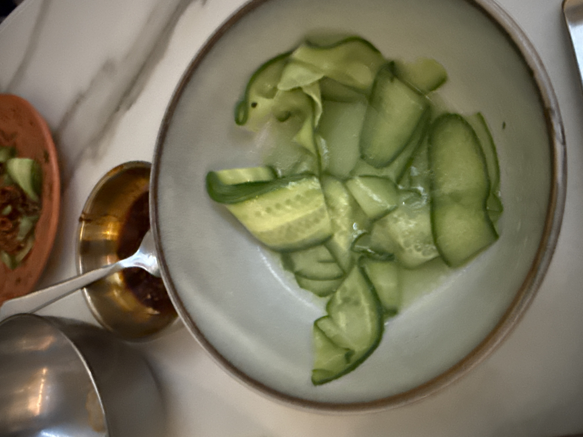 The image shows a top-down view of a table setting in a restaurant, potentially located in Mexico City, Mexico. The table appears to have a marble or similar surface. The primary focus is a shallow, round bowl filled with thin cucumber slices dressed in a light sauce. Adjacent to this bowl, there is a small, golden-colored dish containing a dark sauce, possibly chili oil or a similar condiment. A spoon with a silver finish rests in the sauce dish. To the left of the scene, there is a glimpse of another reddish-brown bowl with what seems to be a salad. A silver-colored serving dish can be seen at the bottom left corner. A metal utensil is present at the top right. The overall scene suggests a shared dining experience or a selection of small plates.