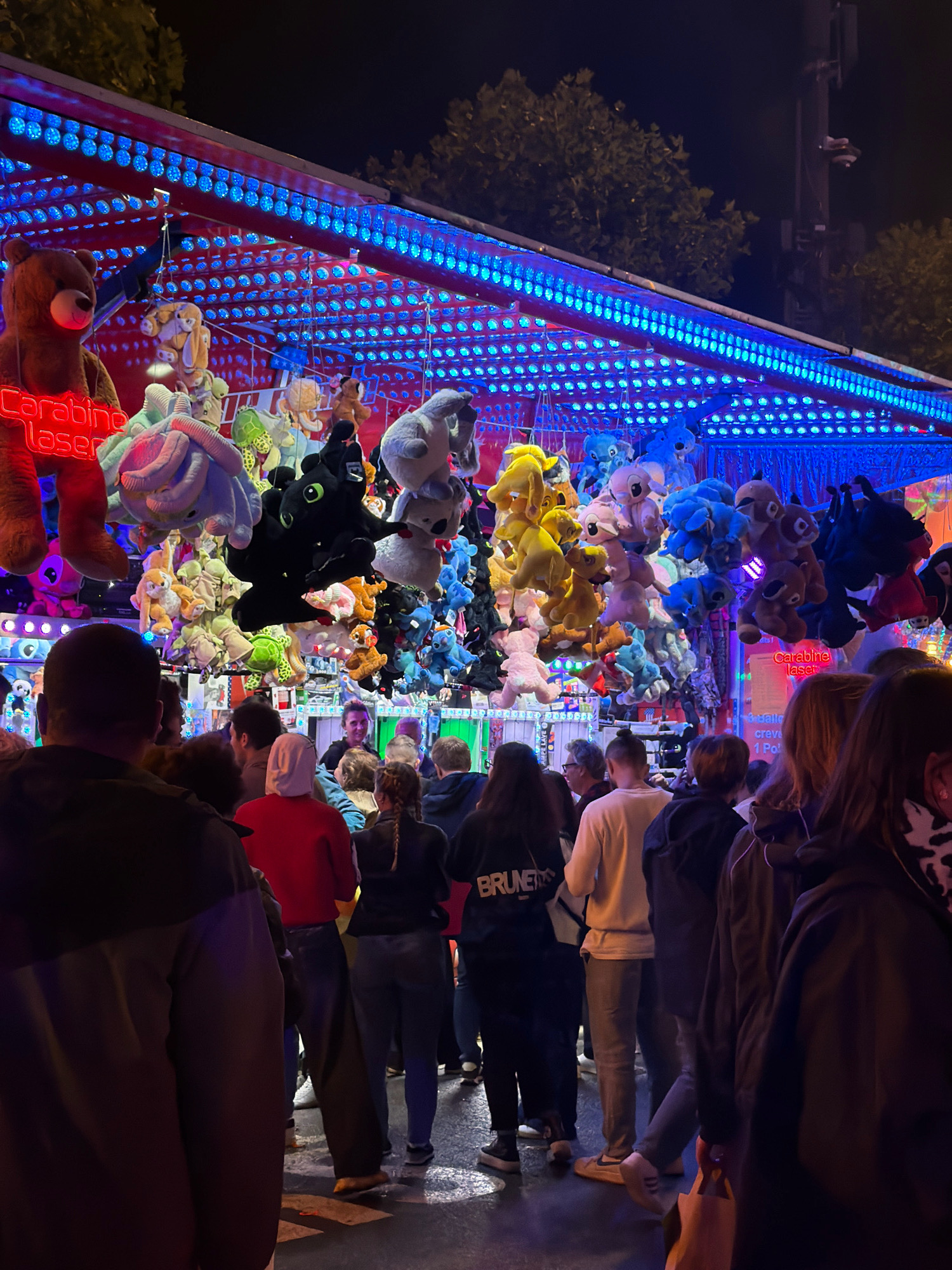 The image depicts a nighttime scene at a fairground, likely during a public event or festival in Luxembourg City. A brightly lit stall, adorned with numerous blue lights, showcases a large assortment of plush toys hanging from above. The toys vary in color and character, including bears, elephants, and other cartoonish figures. In the foreground, a crowd of people stands in front of the stall, some facing the stall and others looking away, suggesting they are queuing or observing the attractions. The ground appears wet, possibly from rain or recent cleaning, reflecting the surrounding lights. A sign with the text "Carabine Laser" is visible on the stall, indicating a potential game or activity. The overall atmosphere is festive and lively, characteristic of a fairground at night.