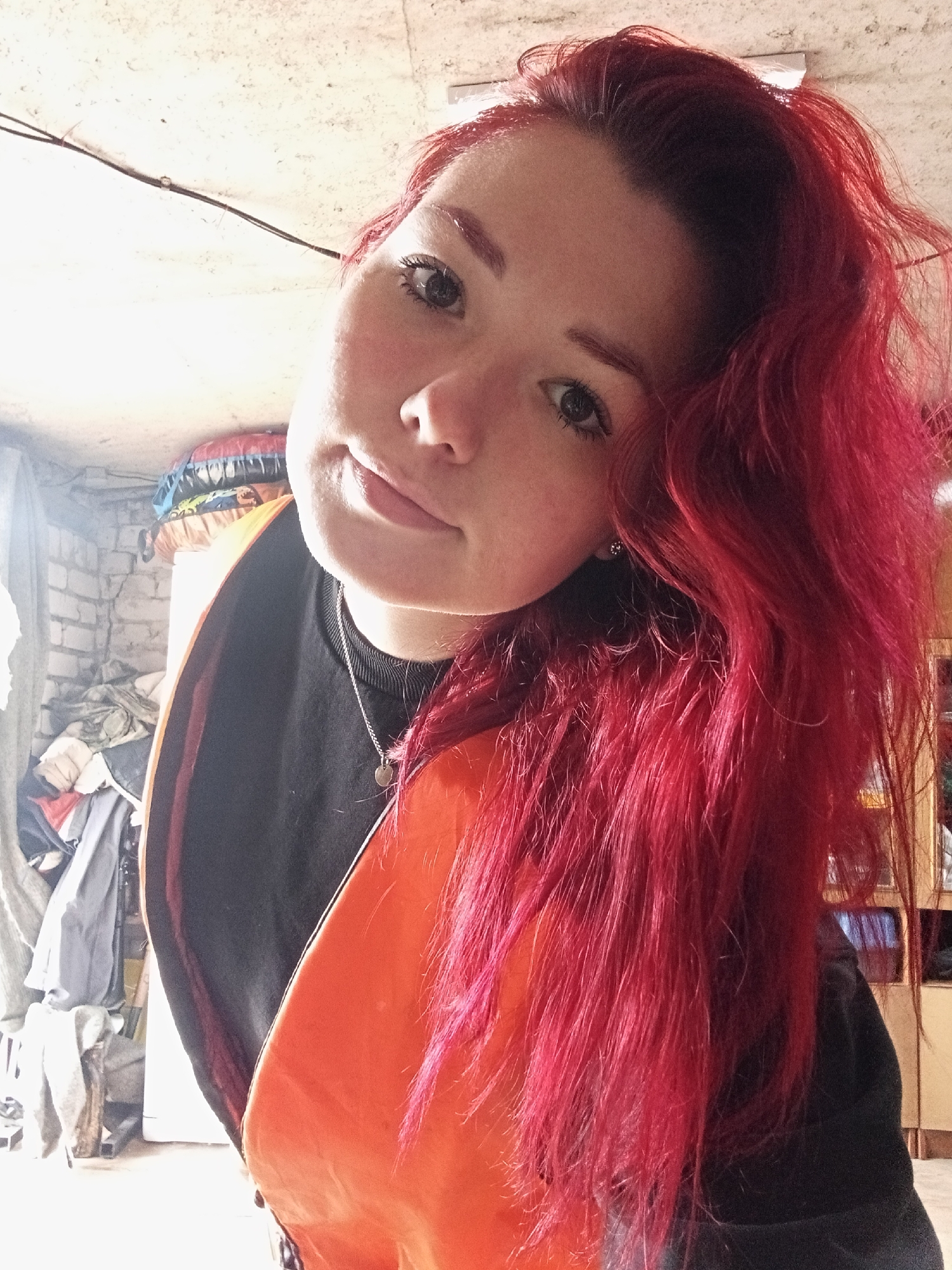 An extreme close-up, low-angle selfie is captured, featuring a young woman with vibrant red hair, styled in loose waves cascading down her right side. She is looking directly at the camera with a subtle smile. Her left eyebrow appears to be tinted red, matching her hair. She is wearing a black crewneck shirt with a small silver pendant necklace. A bright orange fabric is draped over her left shoulder, overlapping with the black shirt.
The background is dimly lit and appears to be an indoor, possibly unfinished space. To the left, a brick wall is visible, with various items of clothing and other objects stacked against it. A window or opening lets in a bright light from the left. The ceiling is textured and shows electrical wiring. To the right of the woman, wooden shelving or furniture is partially visible, with some reflections on its surface. The overall environment suggests a casual, possibly cluttered personal space.