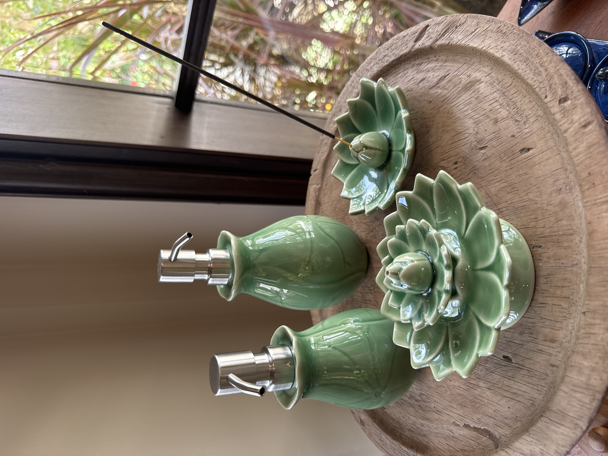 The image shows a collection of decorative items arranged on a weathered wooden tray. Two green, floral-shaped soap dispensers with stainless steel pumps are present. There are also two similarly styled floral incense holders; one is holding a lit incense stick. The tray sits indoors near a window, through which foliage can be seen.
The location is Kuta Selatan, Indonesia.