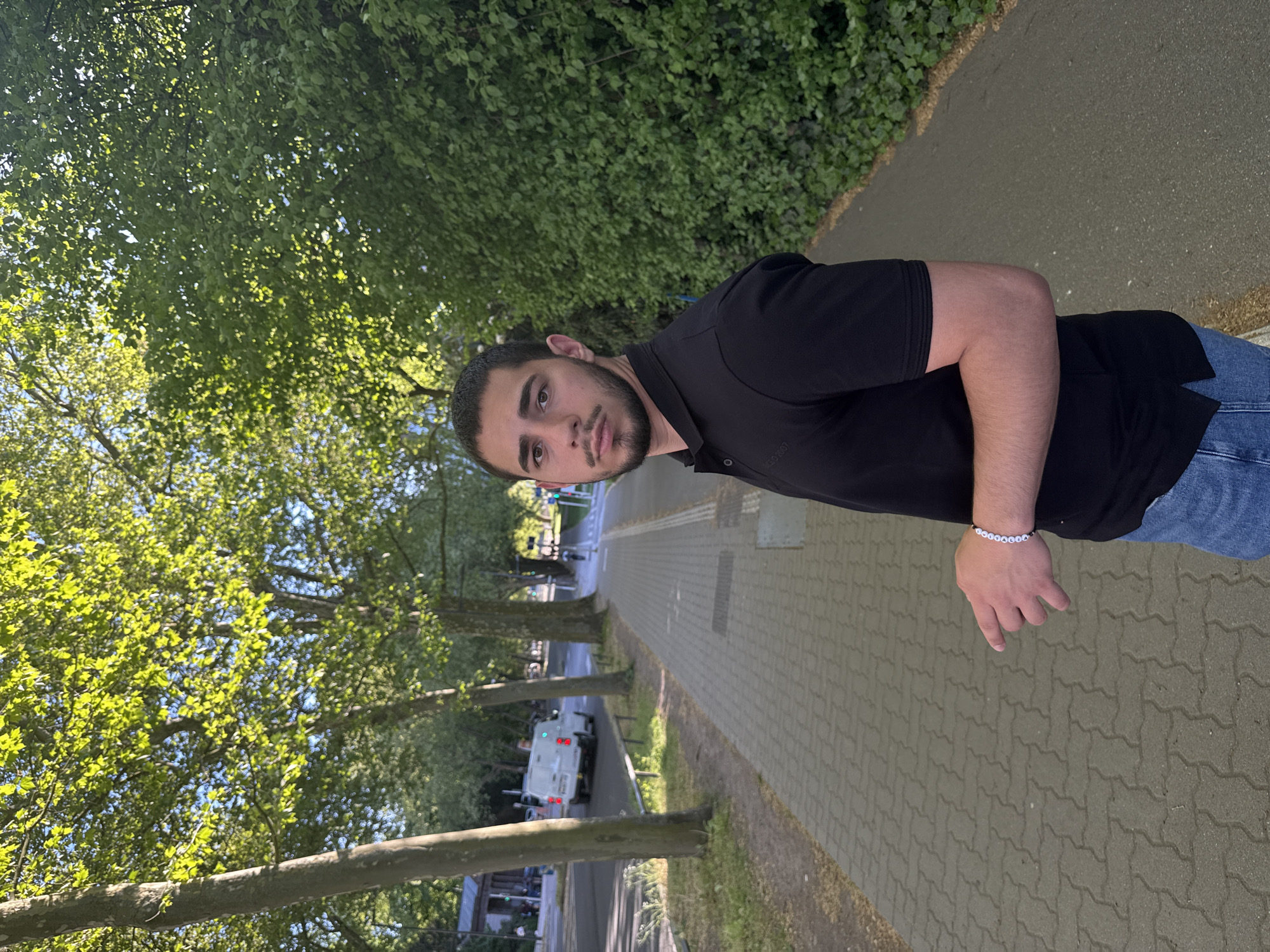 A young man stands on a brick sidewalk under the shade of trees along a road. He is wearing a black polo shirt and jeans. He is looking toward the camera. The road is paved and has a white van or truck parked on the side. There is green foliage on either side of the road and the sidewalk. Based on the appearance of the architecture and foliage, the scene may be within the city of Strasbourg, France.