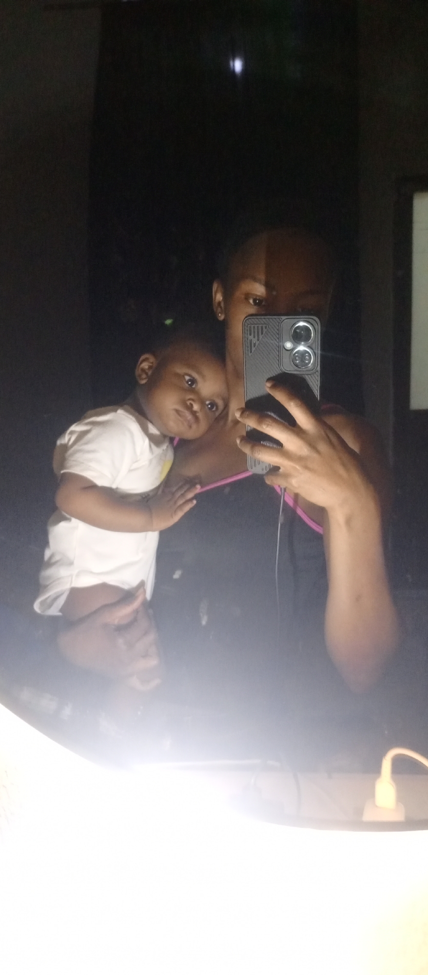 A person is holding a baby while taking a selfie in a mirror. The person is wearing a black tank top with pink straps. The baby is wearing a white onesie. The background is dark. The location is Unknown, Unknown.