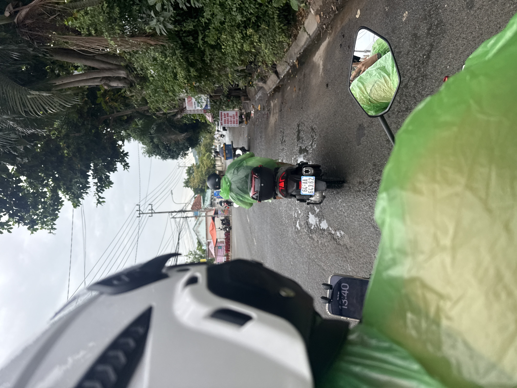 The image captures a first-person perspective from a motorcycle in Nha Trang, Vietnam, on a cloudy day. The rider is wearing a white helmet and a green poncho. In front, another motorcyclist, also in a green poncho, is riding away, their motorcycle plate visible as "64-AA 643.62". The road is wet, indicating recent rain, and appears to be a city street with trees, power lines, and buildings lining the right side. Some small shops or stalls are visible on the left side of the road. A smartphone mounted on the motorcycle displays the time "13:40". The overall scene suggests a typical day of travel on a motorcycle in a Vietnamese city.