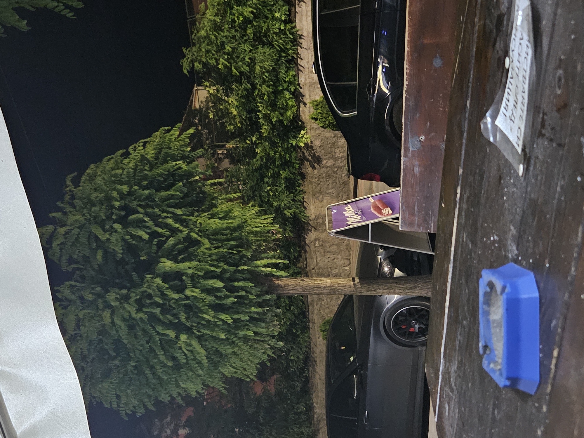 The image depicts a nighttime scene on a wooden table in front of a building. In the foreground, a blue ashtray sits on the table. Behind the table, a street is visible with two parked cars, one dark and one gray, a tree, and a stone wall. A small advertising stand with the "Milka" logo is positioned near the cars. The background shows greenery and parts of buildings. The location is identified as Byala, Bulgaria, suggesting an outdoor seating area of a cafe or restaurant.
