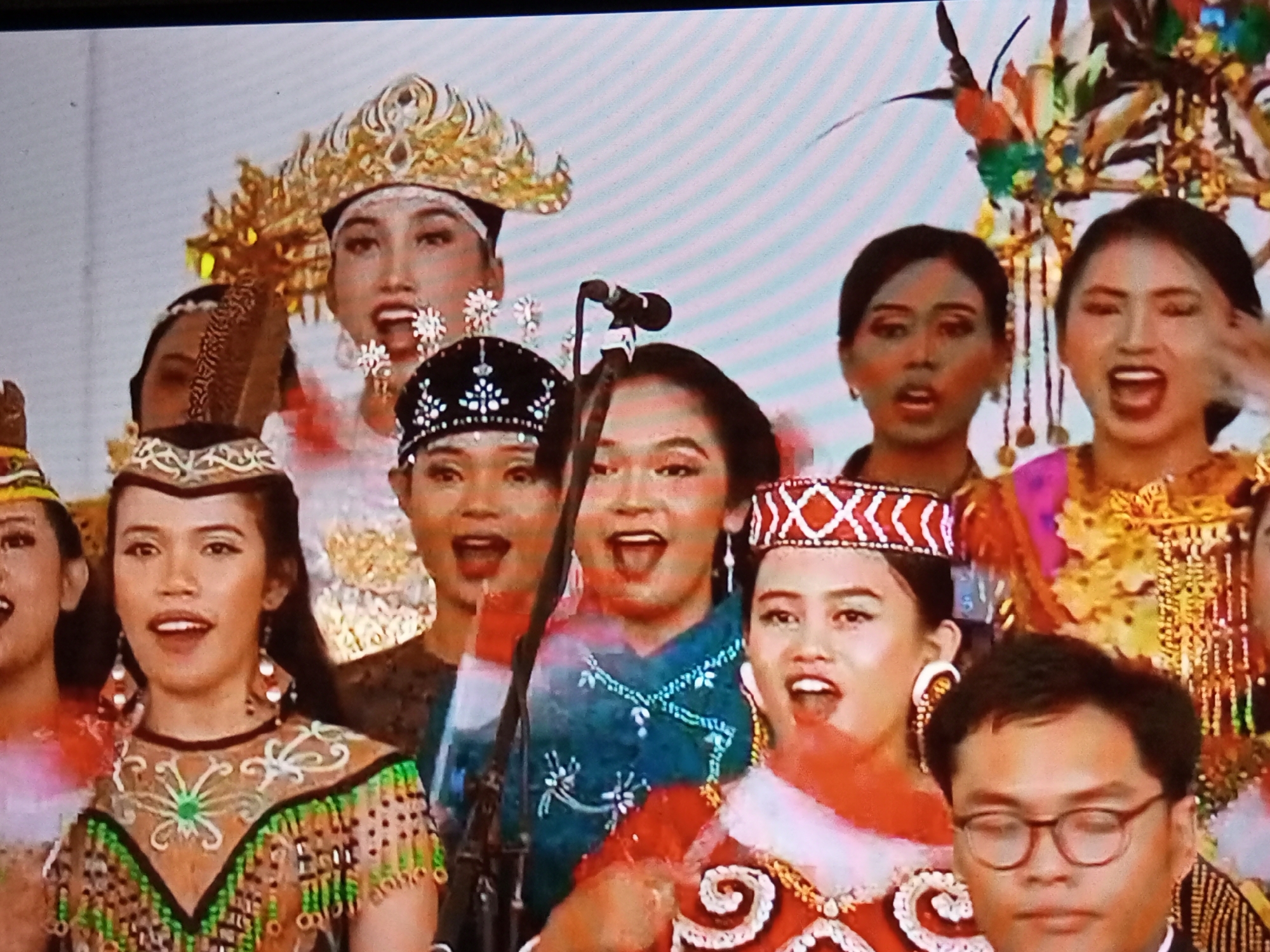 In Banyudono, Indonesia, a group of individuals are performing on a stage. The central figures are women dressed in ornate traditional attire, some wearing elaborate headdresses. They appear to be singing or chanting, as their mouths are open. In the foreground, a man wearing glasses is looking down, possibly attending the performance. The background is a plain, light-colored surface, likely a stage backdrop. The overall scene suggests a cultural performance or festival.