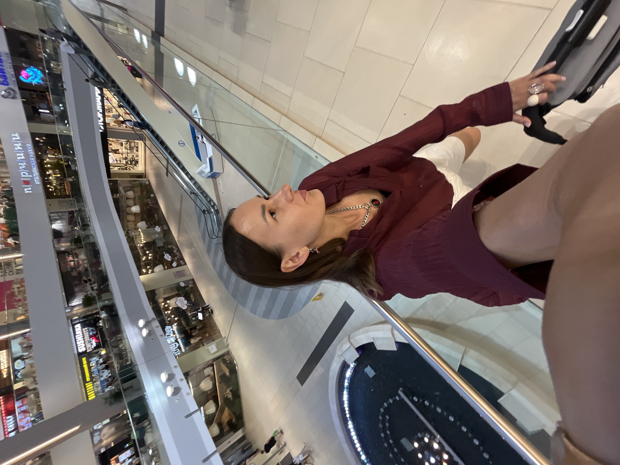 The image shows a woman in a shopping mall, possibly in Sochi, Russia. She is holding a dark gray suitcase with her right hand. She is wearing a dark red collared shirt and white shorts. Her left arm is extended towards the camera, capturing the selfie. In the background, there are escalators, shops, and a fountain. Several people are visible on the escalators and walking in the mall. The mall appears to be multi-leveled with a modern design.