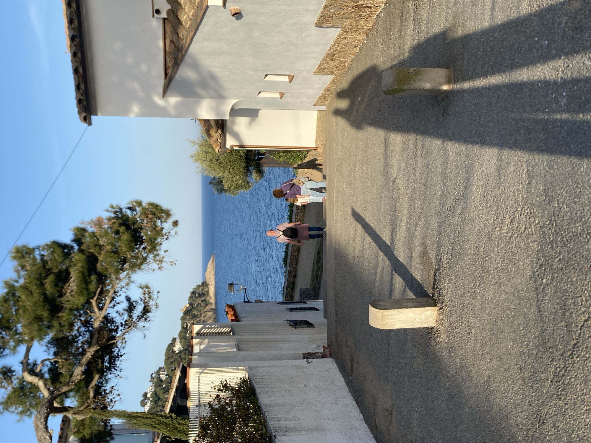 The image captures a street in Calella De Palafrugell, Spain, with a view towards the Mediterranean Sea. Two people, an adult wearing a purple shirt and jeans, and a child, are walking down the street towards the sea. Buildings line both sides of the street, characterized by white facades, tile roofs, and rectangular windows. The street itself is paved and features concrete blocks protruding from the side. Shadows cast by the structures and possibly trees create an interesting contrast on the street surface. In the background, the blue sea is visible along with a small, rocky landmass. A large tree is prominent on the left side of the frame, contributing to the overall atmosphere of the scene.