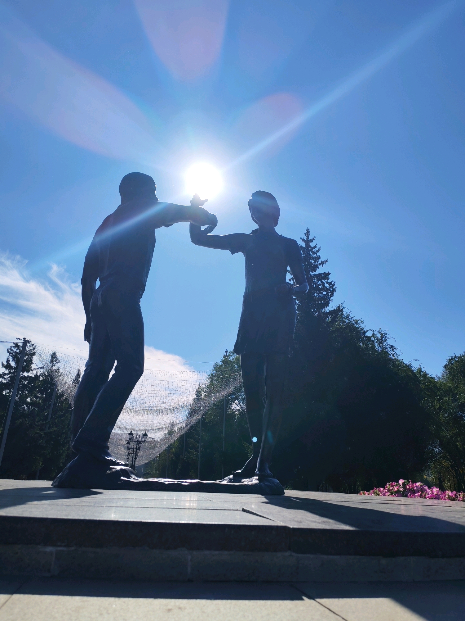 In the city of Unknown, Unknown, a silhouette of a bronze statue depicts a man and woman in a close embrace, facing each other. The man, on the left, is leaning forward with his right arm extended, holding the woman's hand. The woman, on the right, is positioned slightly behind him, her left arm also extended towards him. They are both silhouetted against a bright, hazy sun, which creates lens flare and streaks of light across the bright blue sky. Behind the statues, a line of coniferous trees forms a dark backdrop, with some lighter-colored branches and foliage visible. A string of small, white lights is strung between poles in the middle ground, and a lamppost is visible behind the couple. In the foreground, the statues stand on a tiered granite platform, with the lower part of the platform and the ground visible. To the right, a cluster of pink flowers lies on the platform. The lighting suggests it is daytime, with the sun high in the sky.