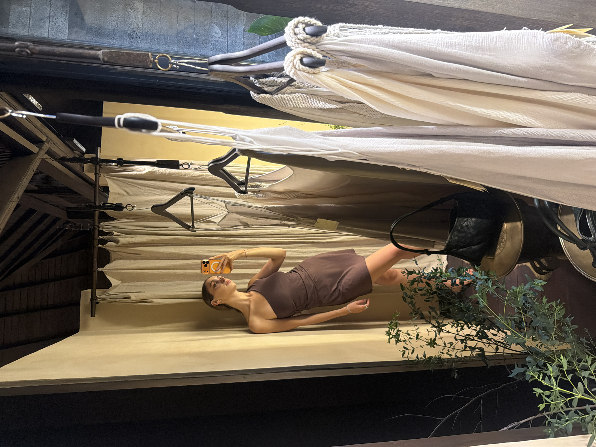 The image captures a woman posing in what appears to be a fashion retail space or showroom, possibly in the Ubud District, Indonesia. The woman, dressed in a form-fitting brown one-shoulder dress, is lying on her side on a light-colored platform or bench, holding a yellow-cased smartphone with a visible MagSafe ring. She is looking upwards, seemingly towards a camera or light source, with a calm expression.
Above her, numerous garments, predominantly in shades of cream, beige, and light grey, are neatly hanging on wooden hangers. Some hangers feature distinctive rope-wrapped details or black foam grips, suggesting boutique-style merchandise. A black woven handbag is visible among the hanging items, resting on a surface. Green leafy branches are positioned to the right of the woman, adding a natural, possibly tropical, accent to the scene.
The background reveals architectural elements, including dark wooden beams or roof structures, and a darker area that could be an outdoor pathway or a shaded part of the building. The lighting is soft and diffused, indicating either an indoor setting or a shaded outdoor area during the daytime. The overall impression is that of a styled photoshoot within a chic, curated retail environment. No visible text is discernible in the image.
