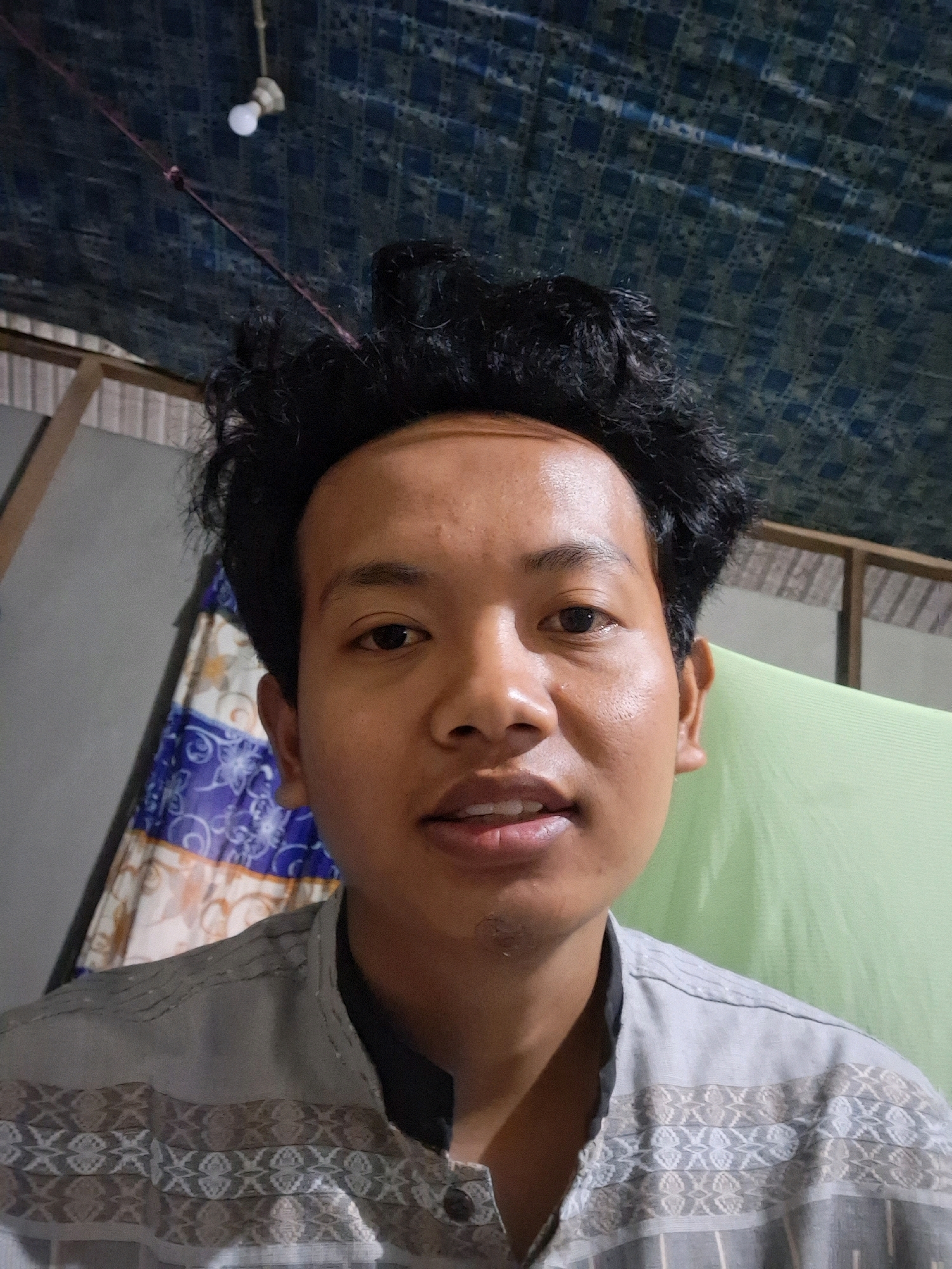 The image shows a close-up selfie of a young man with dark, curly hair. He is wearing a light grey collared shirt with a pattern of alternating geometric shapes and stripes. The background is out of focus, but appears to be an indoor setting with a patterned ceiling in dark blue, a light bulb hanging from a wire, and a light green fabric or curtain to the right. To the left, a portion of a patterned cloth with blue and white designs is visible. The man's expression is neutral, and he is looking directly at the camera. The image quality suggests it was taken with a mobile phone camera in low light conditions. This scene is identified as being in the city of Air Tawar, Indonesia.