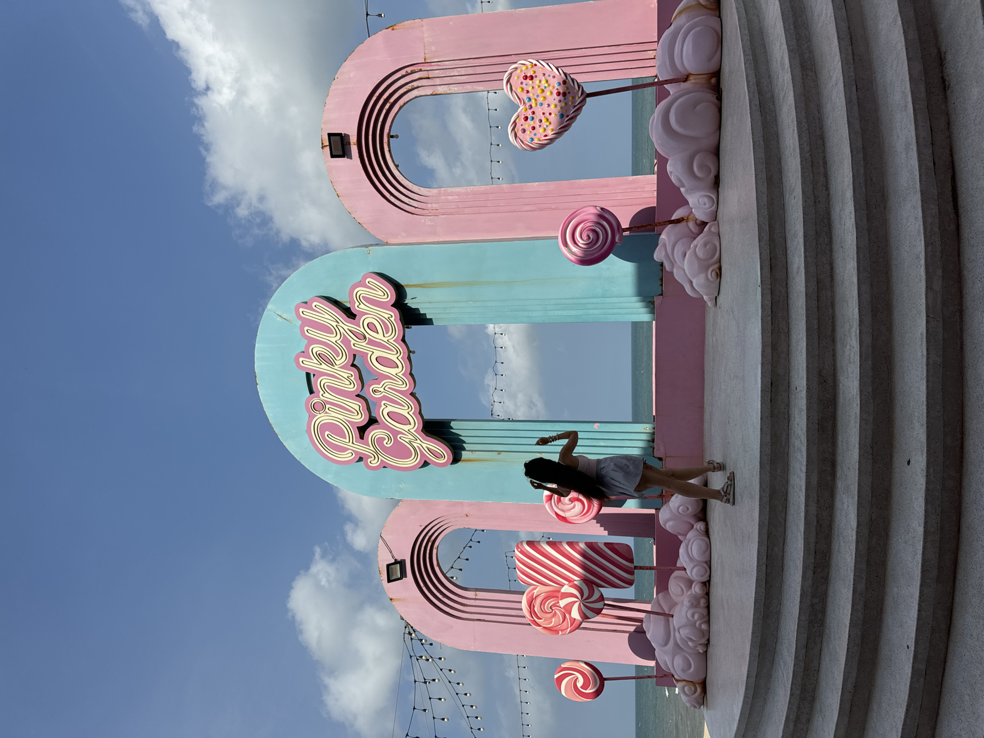 A full shot shows a person from behind, standing on a curved, multi-tiered concrete platform with a bright pink archway structure behind them. The archway is adorned with large, colorful lollipop decorations in various sizes and styles. The center of the archway features a teal panel with "Pinky Garden" written in pink neon letters. The structure is set against a backdrop of the ocean and a bright blue sky with scattered white clouds. The person, dressed in a light-colored tank top and a short skirt, is facing away from the camera, looking out at the ocean. They have long, dark hair and are wearing sandals. The platform itself is made of light gray concrete with a textured surface. The overall scene is vibrant and whimsical, suggesting a theme park or recreational area. The location is in Tien Thanh Ward, Vietnam, specifically at the "Pinky Garden" attraction, known for its candy-themed decor and seaside setting. String lights hang across the top of the archway structure, adding to the festive atmosphere.