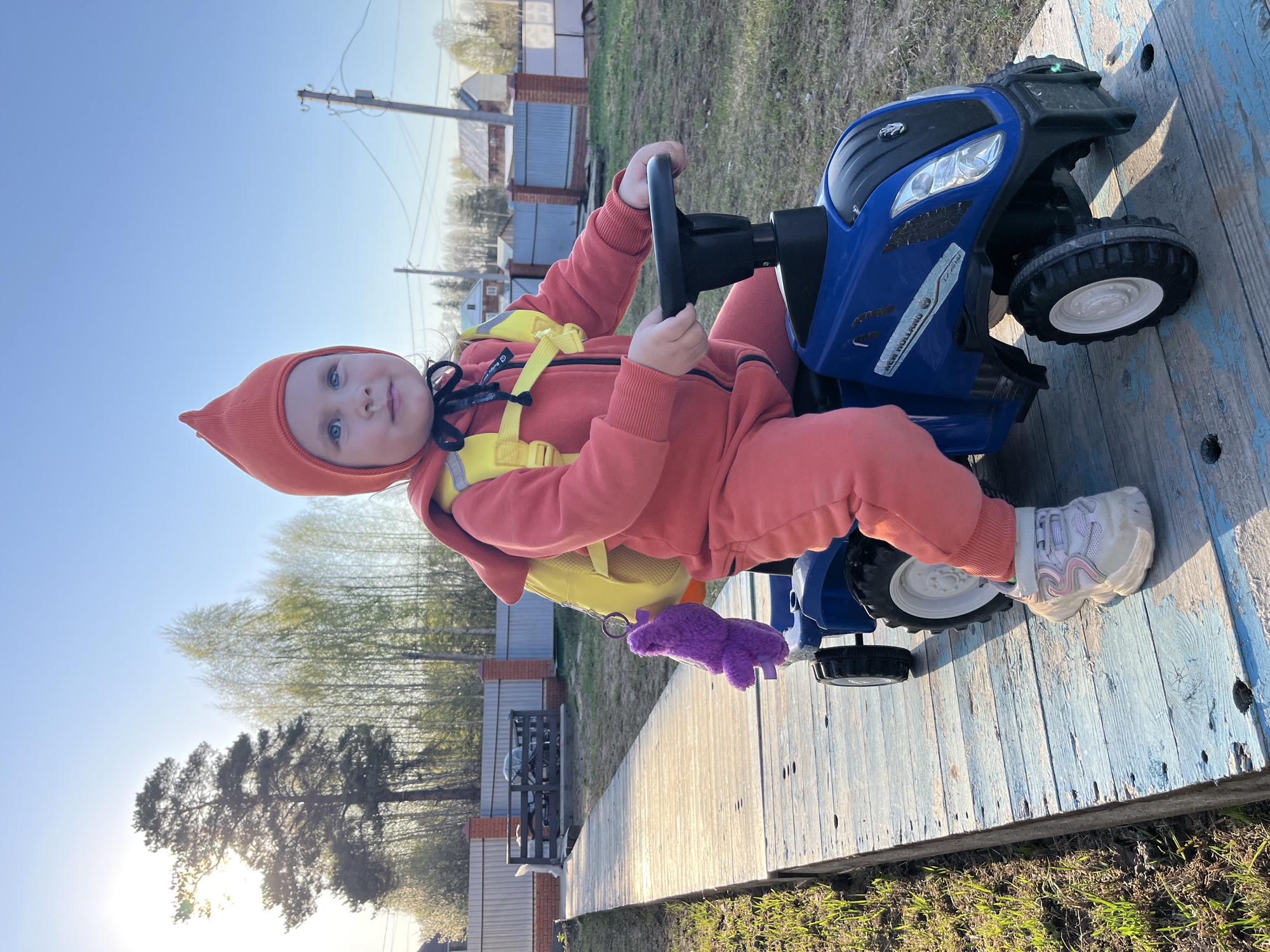 A young child wearing an orange hooded jumpsuit and yellow harness sits on a blue toy tractor on top of a weathered wooden platform. The location is Chaikovsky, Russia, as indicated by the text visible on the New Holland tractor. The platform has a blue-painted surface with visible wood grain and dark patches. The child holds the steering wheel of the tractor with both hands and looks slightly to the left of the viewer. A purple stuffed animal is attached to the harness. The background shows a residential area with brick fences, buildings, and trees. The sky is clear and blue, suggesting a sunny day. The image has been rotated, causing it to appear sideways.