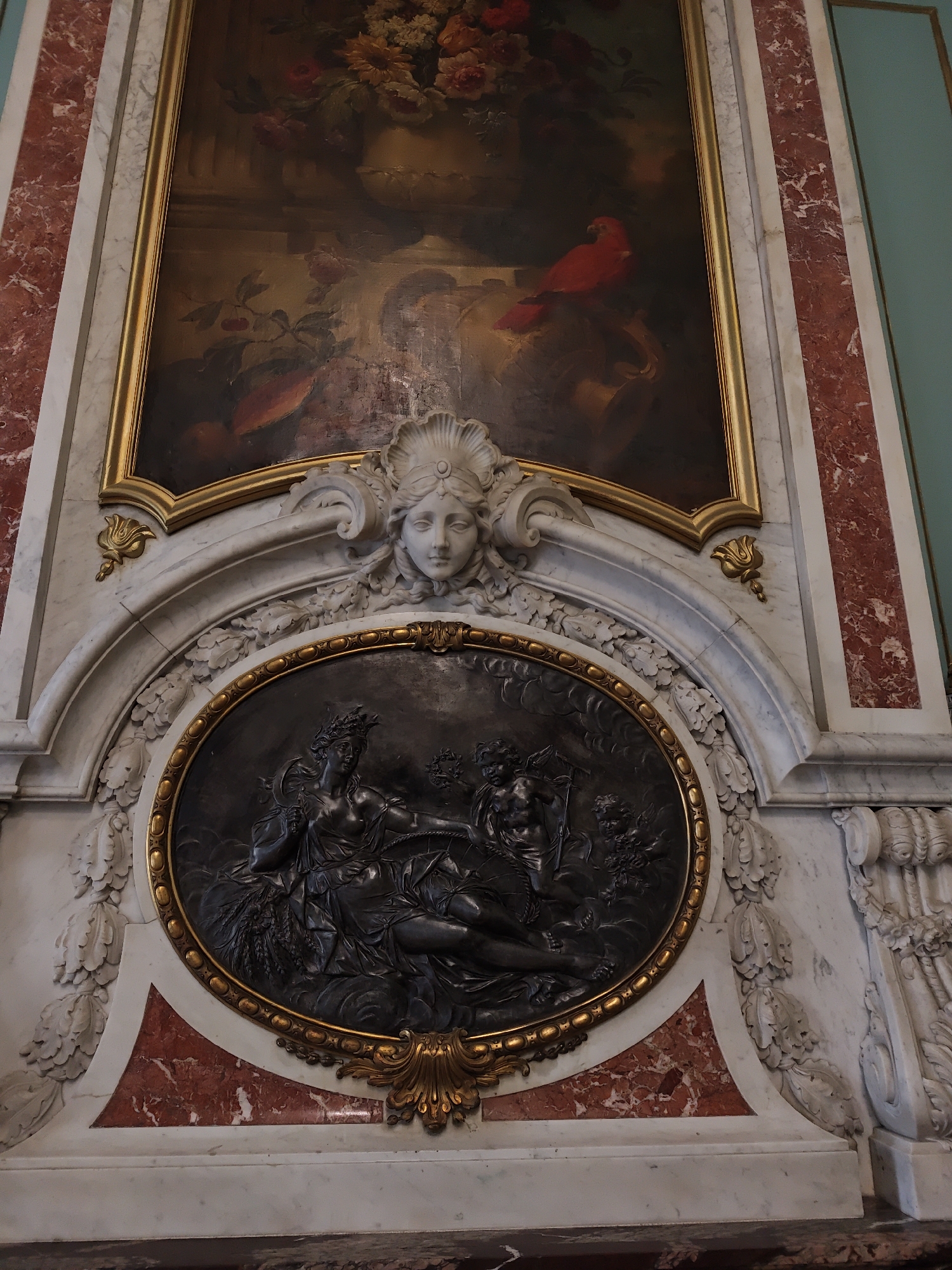 The image shows an ornate fireplace in Lviv, Ukraine, likely within an opulent building or museum. The fireplace features a large, framed painting of a still life with flowers and a red bird. Below the painting, there is a relief sculpture of a woman's face with a seashell headdress, carved into white marble. Further down, an oval relief depicts two figures, possibly mythological, adorned with crowns and holding various symbolic objects, set within a gilded frame. The entire structure is made of marble, with decorative red marble accents and intricate carvings of leaves and floral motifs. The lighting suggests an indoor setting, possibly illuminated by ambient light or artificial sources.
