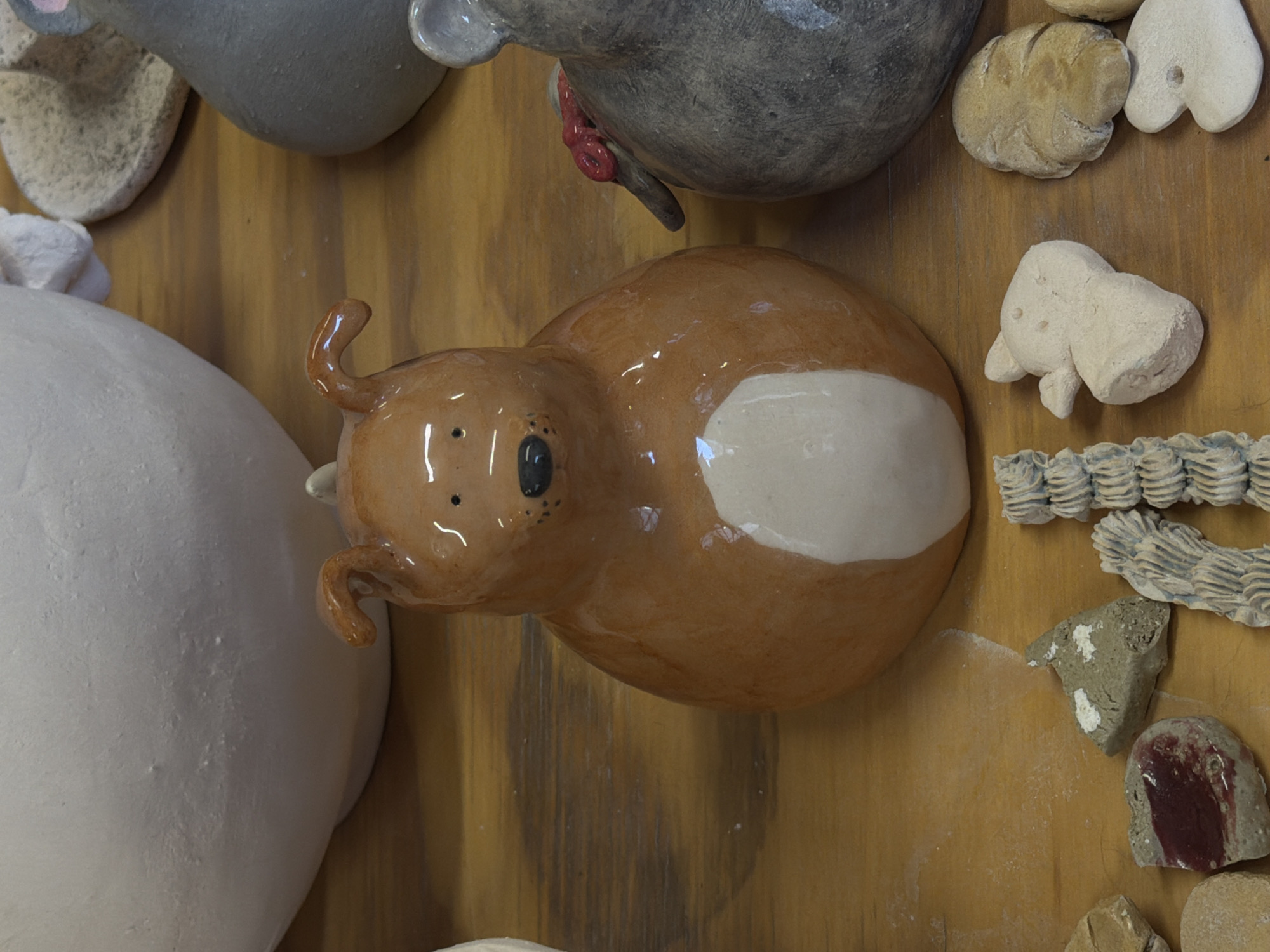 The image displays an overhead view of various ceramic objects arranged on a wooden surface. The primary subject is a glossy, reddish-brown ceramic piece, roughly oval-shaped, resembling a stylized animal with small, curled ears or horns, black dot eyes, and a distinct white patch on its belly. This piece is positioned centrally and slightly to the right, facing towards the left side of the frame.
To its left, a large, smooth, unglazed white ceramic object, appearing as a rounded form, occupies a significant portion of the lower-left corner. Partially visible in the top-left is a gray ceramic object with a textured surface, featuring a small, red, convoluted attachment.
Scattered to the right and bottom of the central brown ceramic piece are several smaller, irregularly shaped ceramic fragments. These include off-white, roughly sculpted pieces, some resembling abstract forms or small animals, and several pieces with a distinct ridged or woven texture in light gray and white tones. There are also irregular, earthy-toned pieces with patches of white and deep red.
The objects are stationary, suggesting they are either displayed, stored, or in a state of drying or post-firing. The background is a warm-toned wooden surface with visible grain, possibly a tabletop or shelf. There is no discernible activity, human presence, specific time of day, or visible text. The setting appears to be an indoor environment, likely a ceramics studio, workshop, or art display area.