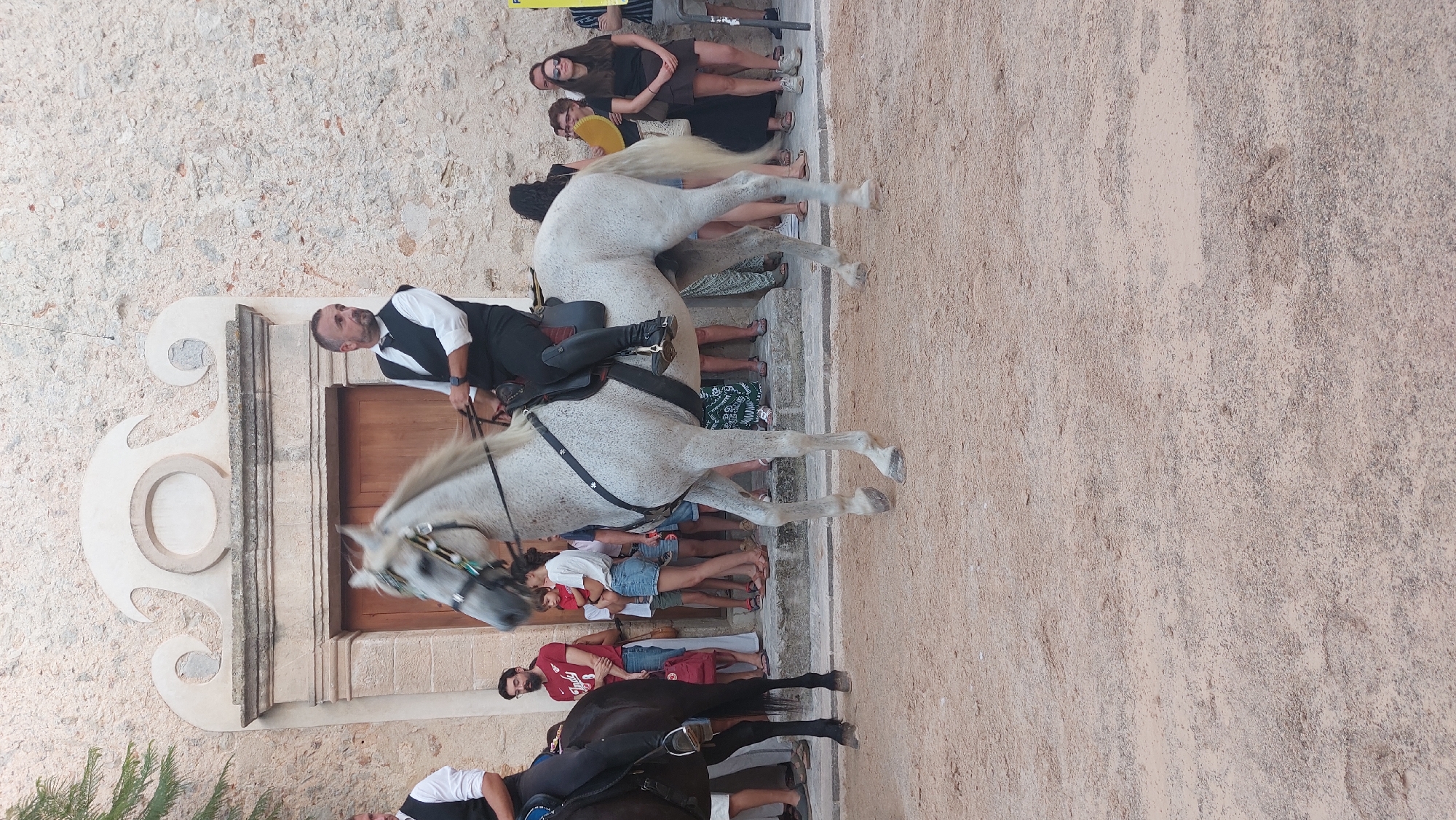 In Costitx, Spain, a man rides a white horse in front of a building with a stone facade and a large wooden door. The man is wearing a dark vest over a white shirt and dark pants tucked into riding boots. He is holding the reins of the horse, which is decorated with a bridle and some colorful ribbons on its head. The horse has a dappled coat. Behind the horse and rider, a crowd of people stands watching. Some of the spectators are young, while others are older. They are dressed in casual clothing, and some are holding bags or fanning themselves. The ground in front of the building is covered in sand. A portion of another horse, black in color, is visible to the left of the white horse.