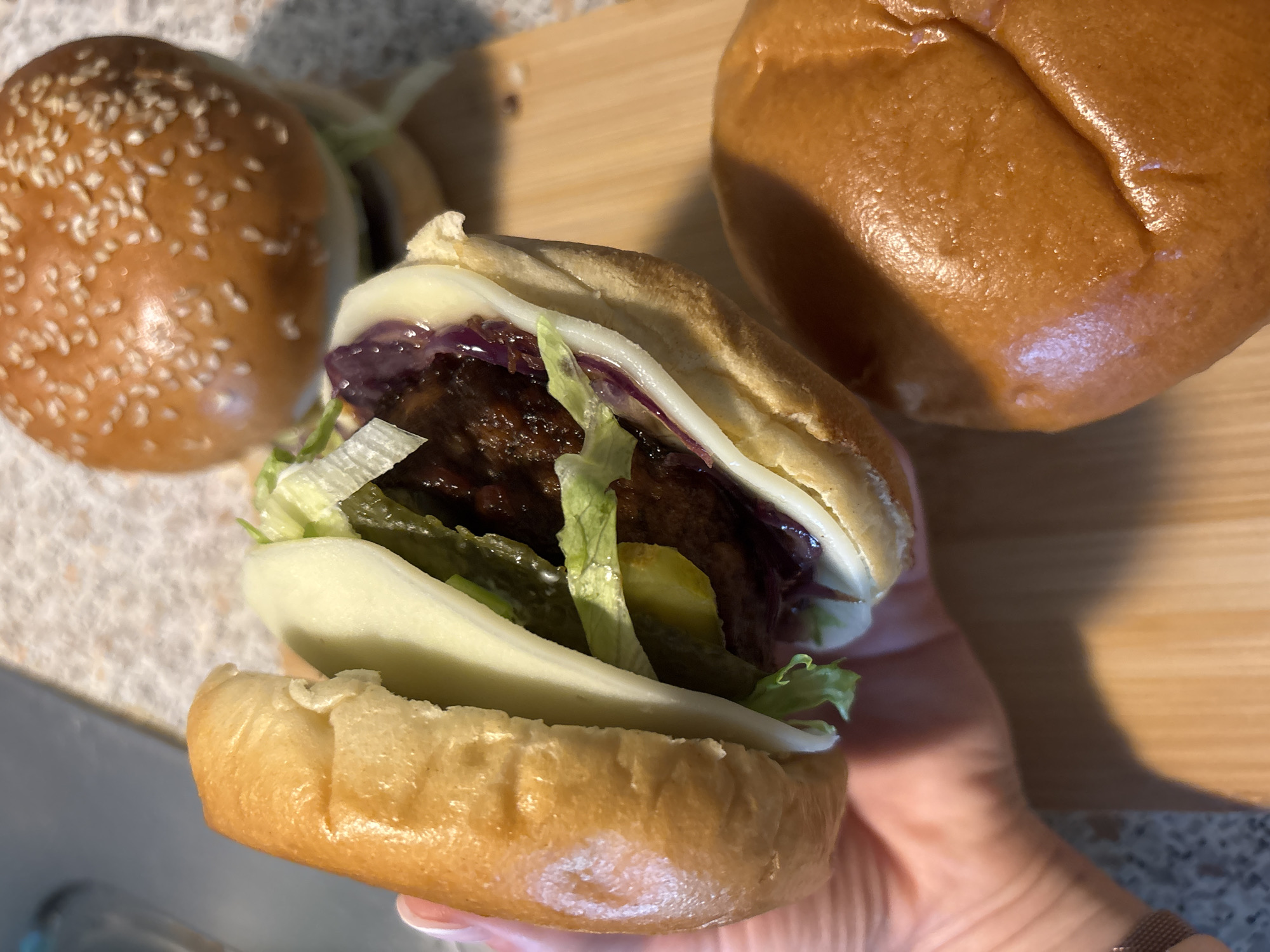 In this overhead shot, a hand holds a partially assembled hamburger. The hamburger has a glossy bun, a dark meat patty, lettuce, pickles, cheese, and thinly sliced red onions. Two other hamburger buns, one sprinkled with sesame seeds, are visible in the background, suggesting a meal preparation or gathering. The scene is set on a wooden surface, likely a countertop or table. The lighting suggests it is daytime, possibly indoors, given the lack of harsh shadows and the general brightness. The image does not contain any visible text. The overall impression is casual and domestic. The location context provided is Wörth am Main, Germany.