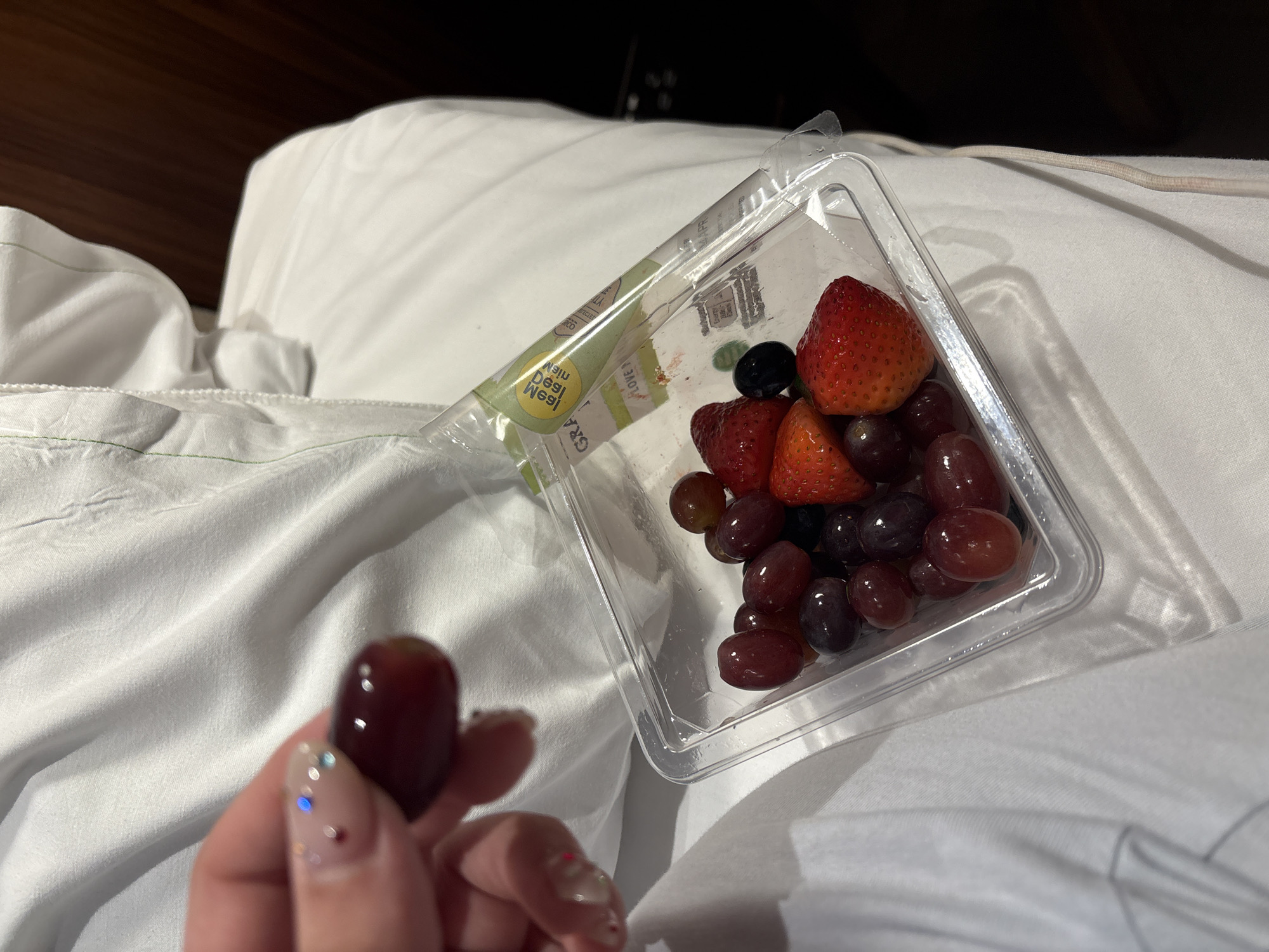 The image shows a clear plastic container of fruit on a white bedsheet. The fruit includes strawberries, blueberries, and red grapes. A hand holds a single red grape in the foreground. The background shows a dark brown wooden headboard. The location is not identifiable as London, United Kingdom.