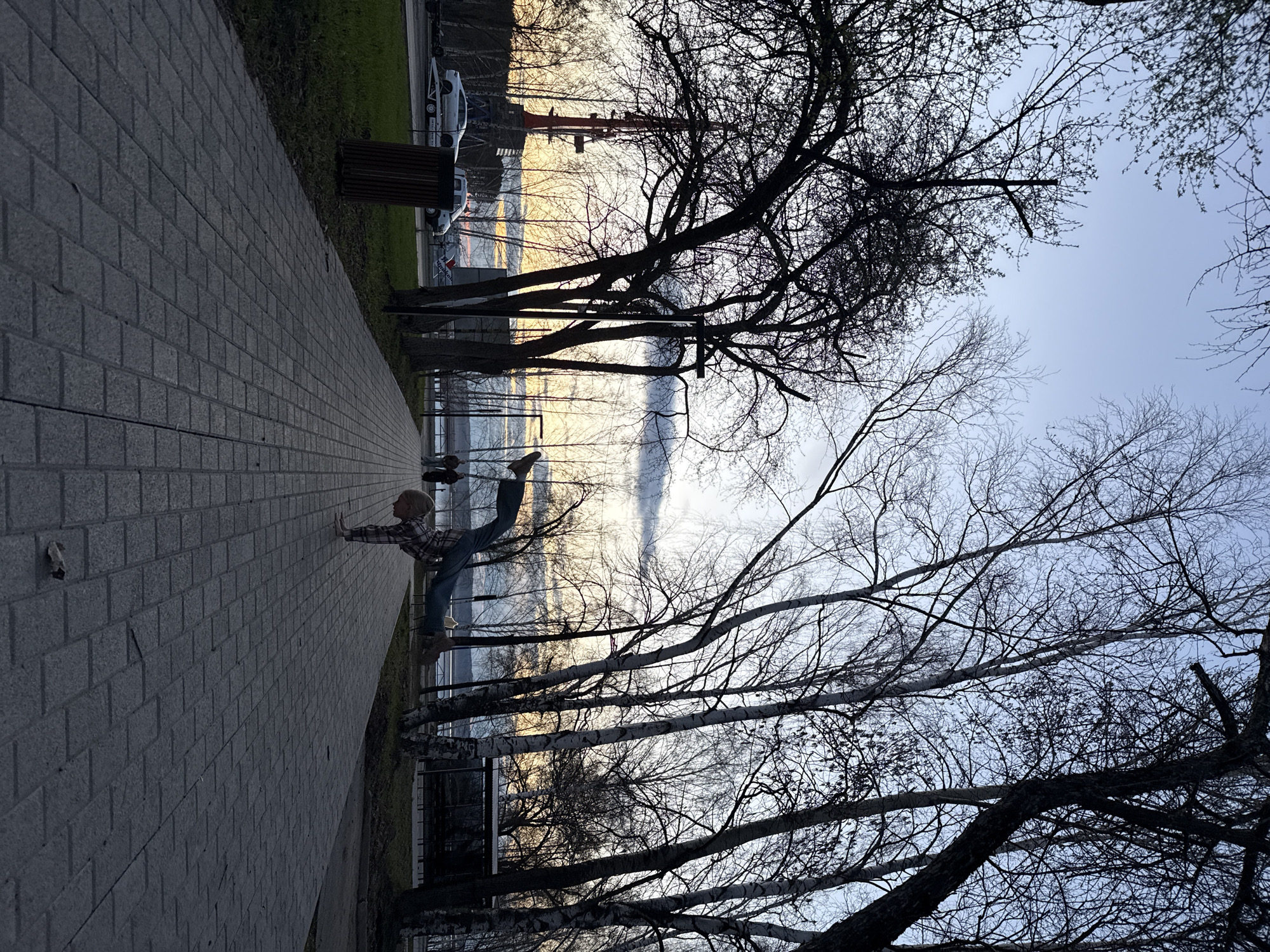 The image, when viewed upright, depicts an outdoor scene along a paved promenade beside a large body of water in Chaikovsky, Russia. The time of day appears to be late afternoon or early evening, characterized by soft, muted light and a pale blue-grey sky that reflects on the water.
The central subject is a young child, dressed in a plaid shirt, blue jeans, and light-colored shoes, with short, light-toned hair. The child is captured mid-action, striking a dynamic balance pose with one leg extended upwards behind them and arms outstretched for stability. This activity suggests playfulness, concentration, or a gymnastic maneuver.
The foreground features a wide paved walkway constructed from grey rectangular pavers, leading into the distance. To the right of the pathway, numerous trees with bare branches stand, silhouetted against the sky and water, indicating a season of late autumn, winter, or early spring. Some of these trees exhibit light-colored bark, possibly birches. Beyond the trees, a vast body of water, likely a lake or river, stretches out, reflecting the sky's soft hues. In the mid-ground, a road runs parallel to the pathway, with several cars, including two white ones, parked on a grassy strip. A dark wooden bench is also visible near the parked vehicles. No discernable text is visible anywhere in the image.