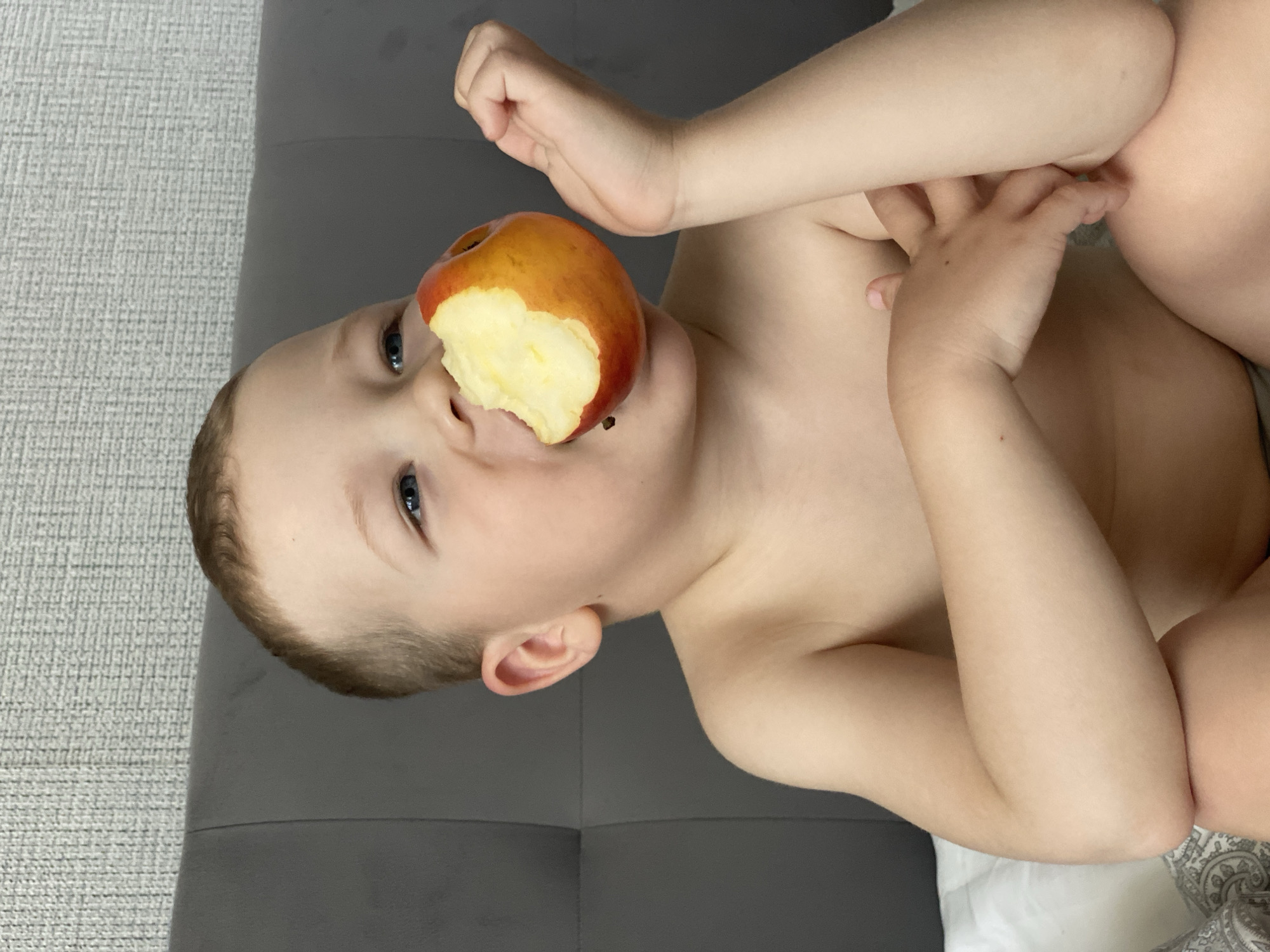 This media file shows a close-up, sideways shot of a young child, likely a toddler, with a partially eaten apple held to their mouth. The child has fair skin, short brown hair, and blue eyes. One arm is raised, touching their face, while the other arm is bent, with fingers tucked near their chest. The child appears to be topless. The background consists of a textured gray surface and a darker gray padded surface, possibly furniture. The lighting suggests an indoor setting, potentially during the daytime. The child's expression is neutral. There is no visible text in the image. The provided location context is Sochi, Russia, but there are no specific visual cues in the image to confirm this location.