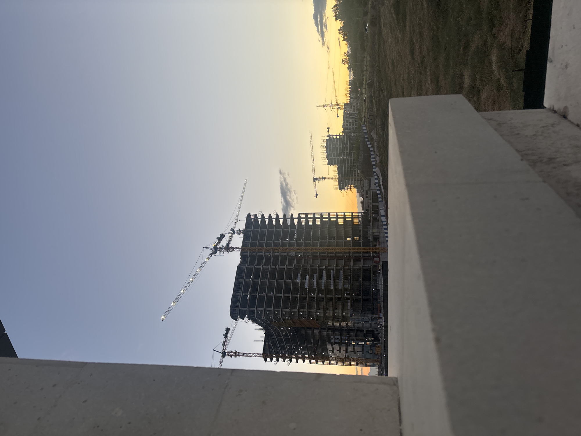 The image captures a panoramic view of a construction site during either sunrise or sunset, evidenced by the gradient sky transitioning from a warm, golden hue at the horizon to a cooler, pale blue higher up. In the foreground, concrete ledges or walls frame the scene from the bottom and right, suggesting the vantage point is from another structure or elevated platform.
The central focus is a large, multi-story building under construction, its skeletal steel and concrete framework clearly visible. Several tower cranes are actively present, two large ones attached directly to this main building, and numerous smaller cranes can be seen in the distance, indicating widespread development. The building's structure shows multiple floors with exposed beams and columns, and some areas appear to have cladding or glass installed, reflecting the warm light of the sky.
Beyond the main construction, the middle ground features a stretch of undeveloped, grassy or barren land. Further in the distance, more buildings under construction are silhouetted against the bright horizon, reinforcing the scale of the ongoing development. The sky is largely clear, with a few wispy clouds scattered across the upper portion. The overall scene suggests significant urban expansion in Yerevan, Armenia, captured at a serene time of day. No visible text is present in the image.