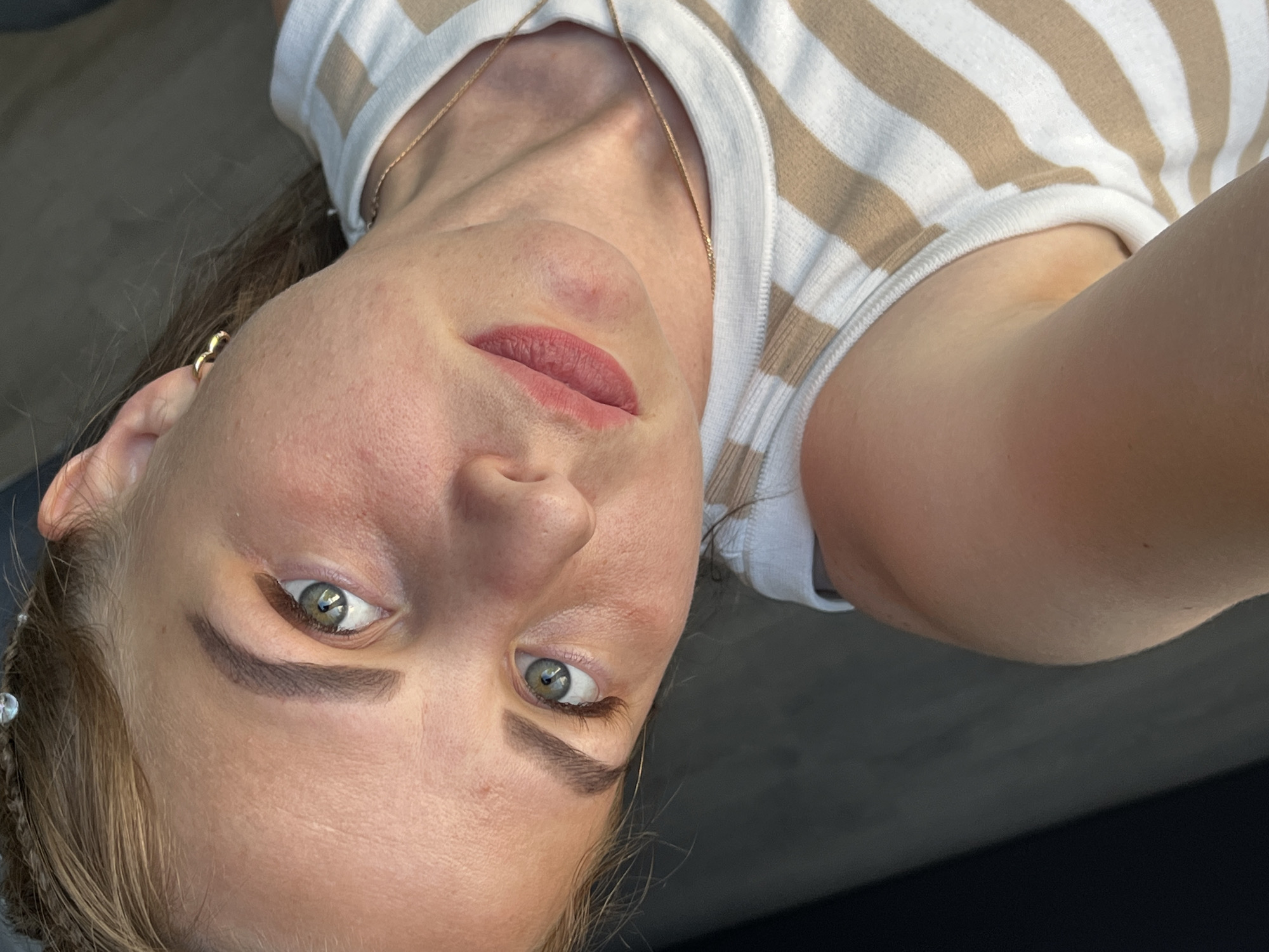 A close-up, angled selfie shows a woman indoors. She has fair skin, light brown hair styled with small braids near her forehead, and green eyes. She wears a white and tan striped tank top and a gold necklace. A small gold earring is visible in her left ear. Her lips are a muted pink color. The background is a dark, indistinct surface, possibly a piece of furniture. There is no evidence suggesting the location is Izhevsk, Russia.