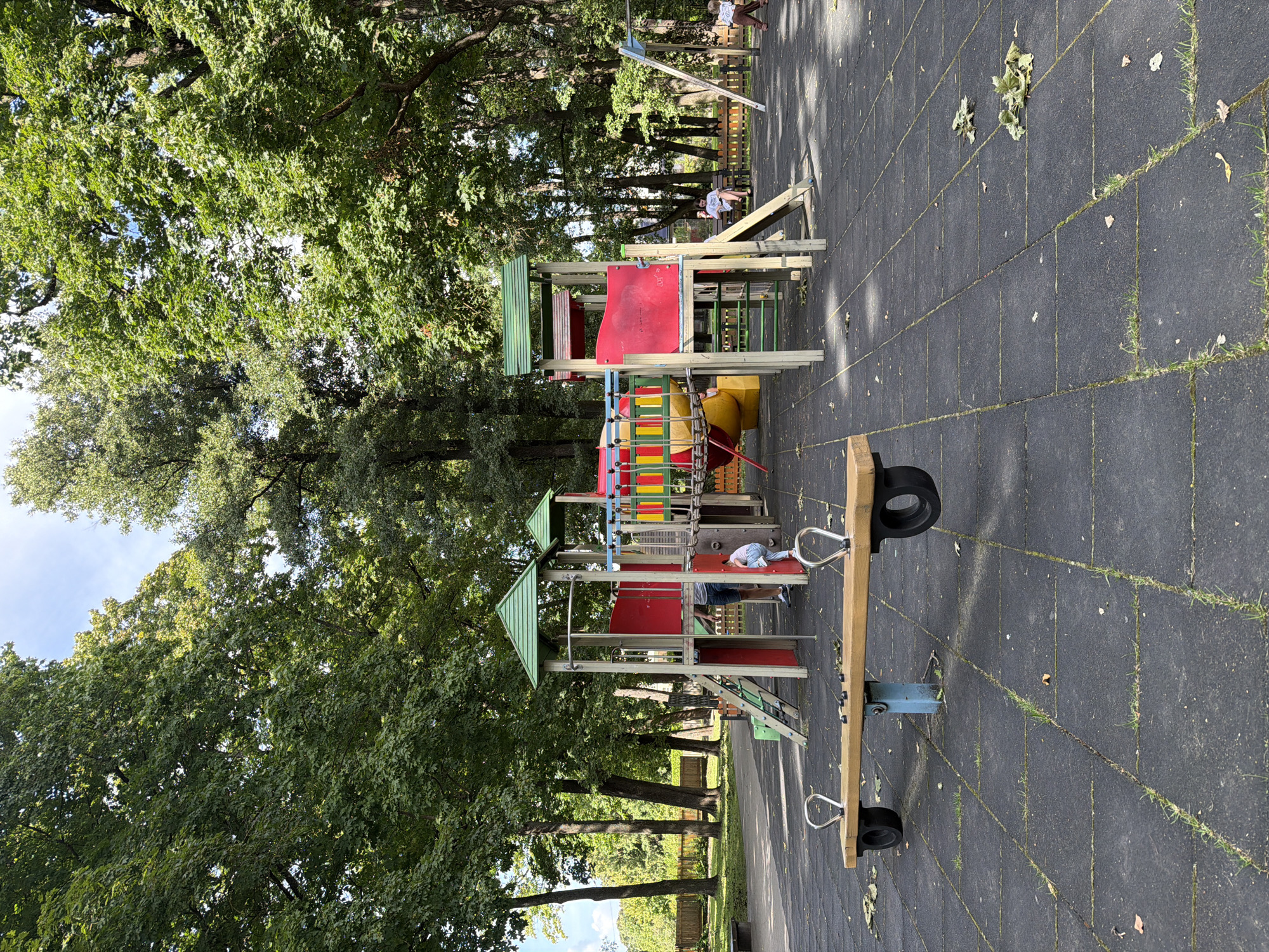 The image depicts an outdoor playground in Riga, Latvia, with a modern, multi-level play structure at its center. The structure is primarily red and yellow with green roofs and features various climbing elements, slides, and bridges. In the foreground, a wooden seesaw with rubber wheels is visible. The ground is covered in dark, interlocking rubber tiles with some patches of grass growing through. The playground is surrounded by lush green trees, indicating a park-like setting. Several people are present; a child is climbing on the play structure, while two adults are seated on a bench in the background. Another child is running near a swing set on the right side of the frame. The sky is partly cloudy.